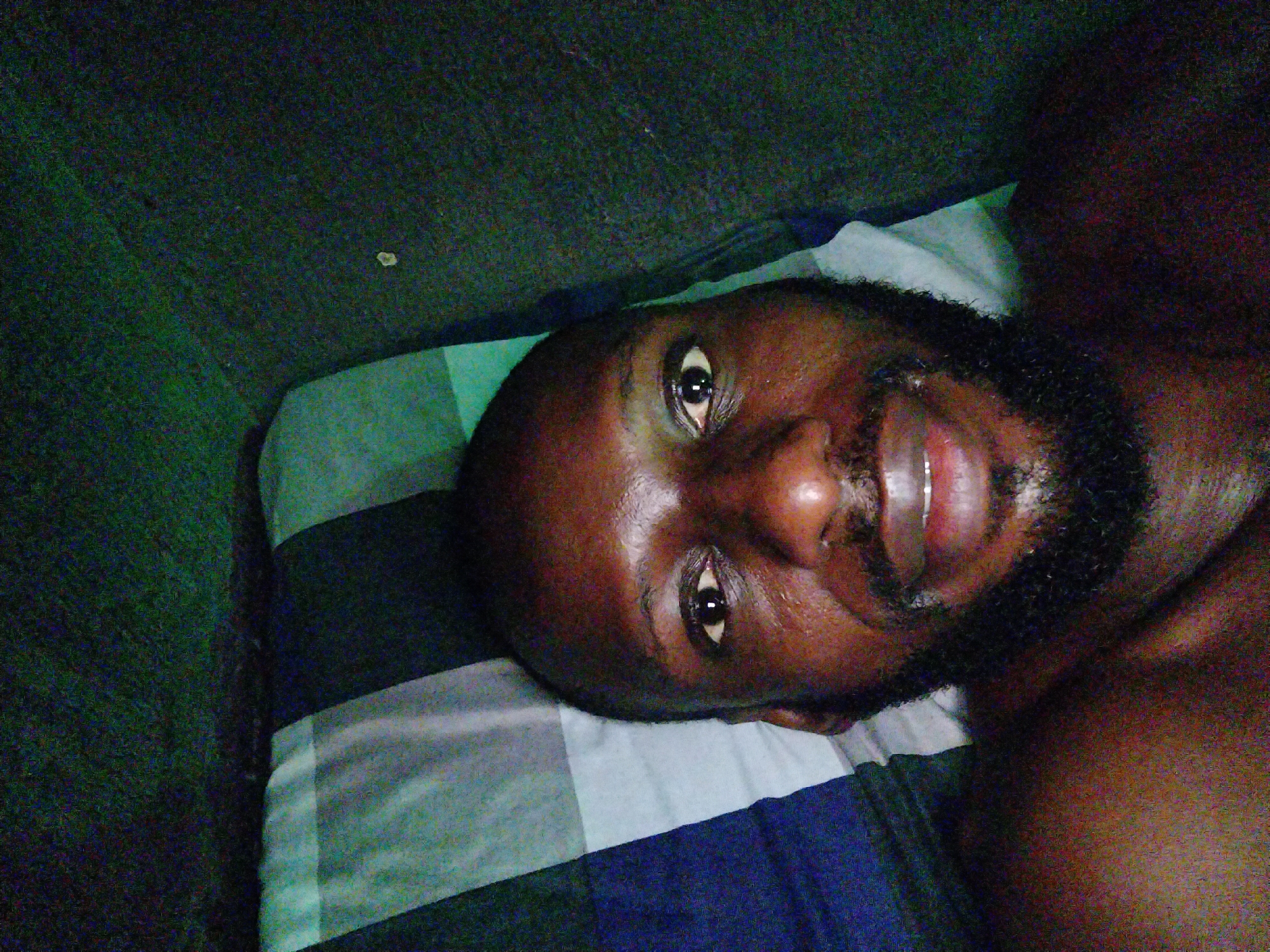 An African man with dark skin and a close-cropped beard lies on a pillow with a blue, white, and green striped pattern. He is looking directly into the camera with his mouth slightly open. The man is indoors, possibly in a bedroom or sleeping space, with a dark green surface visible in the background. It's impossible to pinpoint the exact location in Uyo, Nigeria without more context.