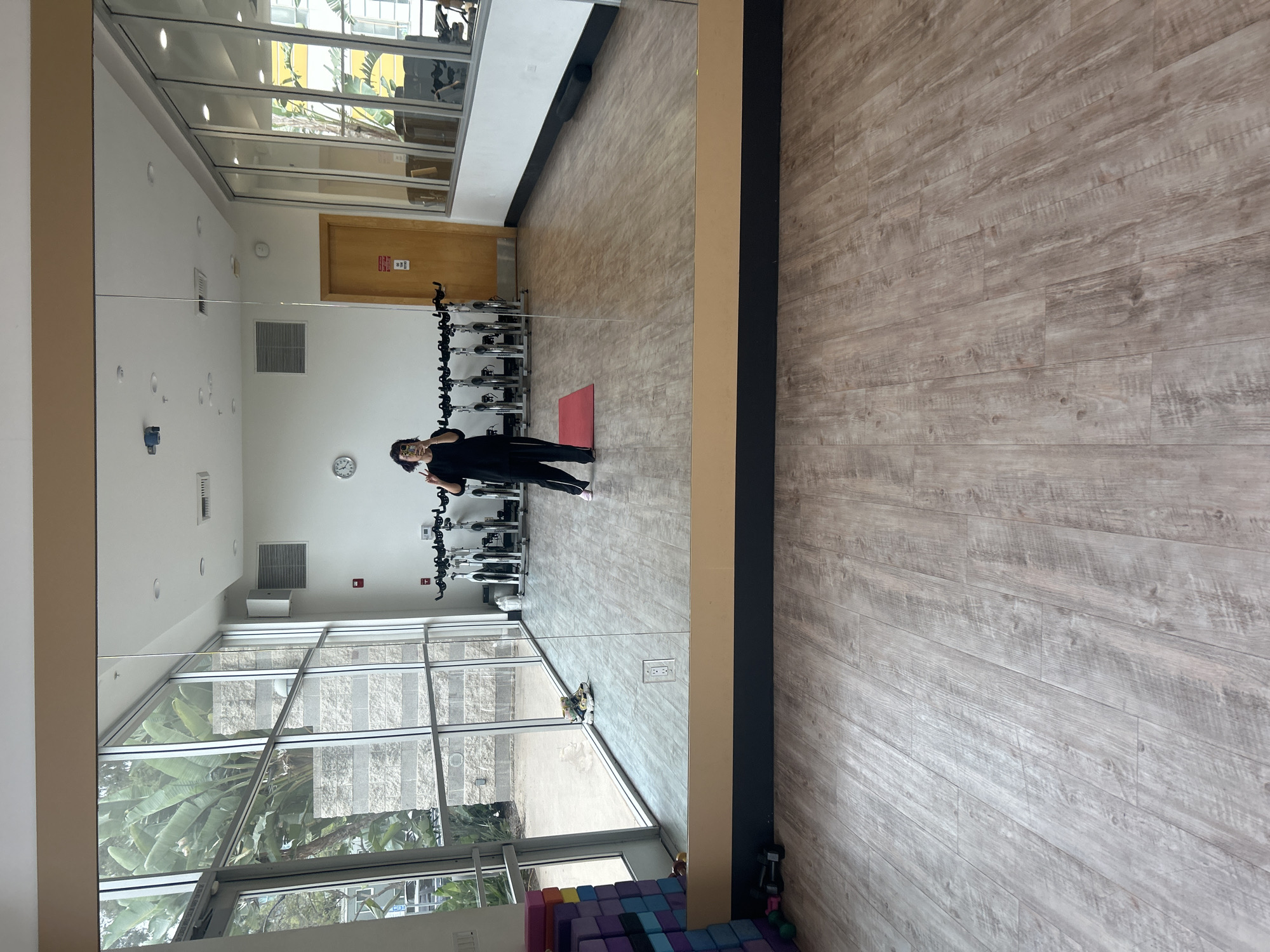 The image captures a mirrored view of a fitness studio in Los Angeles, California. The mirror reflects a woman dressed in black athletic wear posing near a row of stationary bikes and a red yoga mat. The studio has large windows showing exterior greenery, indicating a natural, urban environment. Walls are painted white, and the floor is covered in light brown, wood-patterned flooring. Near the bottom of the mirror, there are yoga blocks and a set of weights. The door is painted in a light brown color. The image is framed to emphasize the mirrored reflection of the scene.
