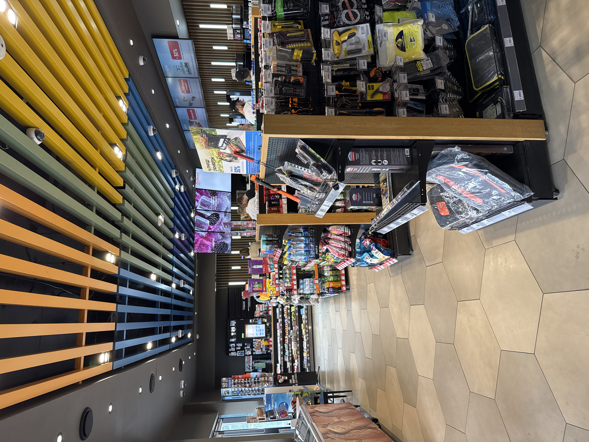 The image depicts the interior of a convenience store, likely a gas station minimarket, with shelves stocked with various goods. The ceiling features a modern design with colorful slats in orange, blue, and yellow, interspersed with lighting fixtures and security cameras. The floor is tiled with a hexagonal pattern in shades of beige and grey.
In the foreground, to the right, a display rack holds tools, including screwdrivers, pliers, and other hardware items, along with cleaning supplies like microfiber cloths. A price tag of "1490p" is visible on one item. To the left, a refrigerated display case is partially visible, along with a black and orange item wrapped in plastic, possibly a car accessory or tool.
Further back, shelves are packed with beverages, snacks, and other convenience items. A counter area is visible with staff members serving customers. Promotional posters are displayed above the counter, one advertising a discount of "50%". The overall scene suggests a well-lit and organized retail environment. The mention of "Sokolov Ruchey, Russia" is not directly derivable from the image itself, but if this context is provided, it would indicate the geographical location of this store.