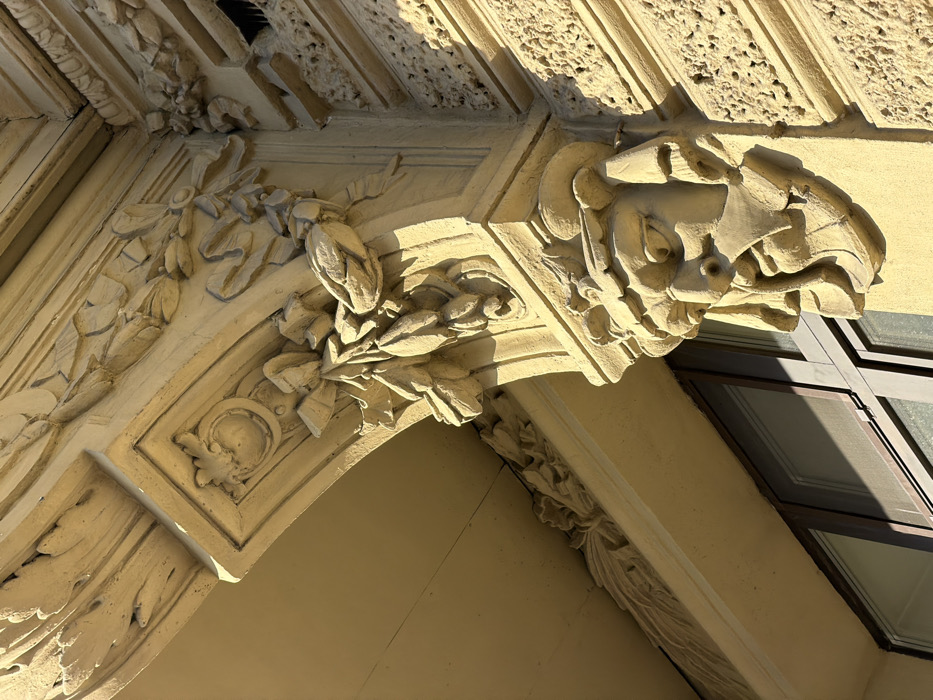 The image displays an ornate architectural detail on the facade of the Singer House (Dom Knigi), located at Nevsky Prospect 28 in Saint Petersburg, Russia. The central element is a pale yellow-beige stone mascaron, depicting a bearded male face with an open mouth and furrowed brow, positioned on a building corner. Above the mascaron, a large projecting console (corbel) features intricate bas-relief carvings of wreaths, garlands, and ribbons, with a decorative circular medallion visible on its underside. To the right of the console, the building wall exhibits a textured, rusticated finish. Below the mascaron and console, the dark frame and reflective glass of a window are partially visible. Strong sunlight from the upper left illuminates the architectural elements, casting sharp shadows that enhance their three-dimensional relief. No people or actions are present in the scene.