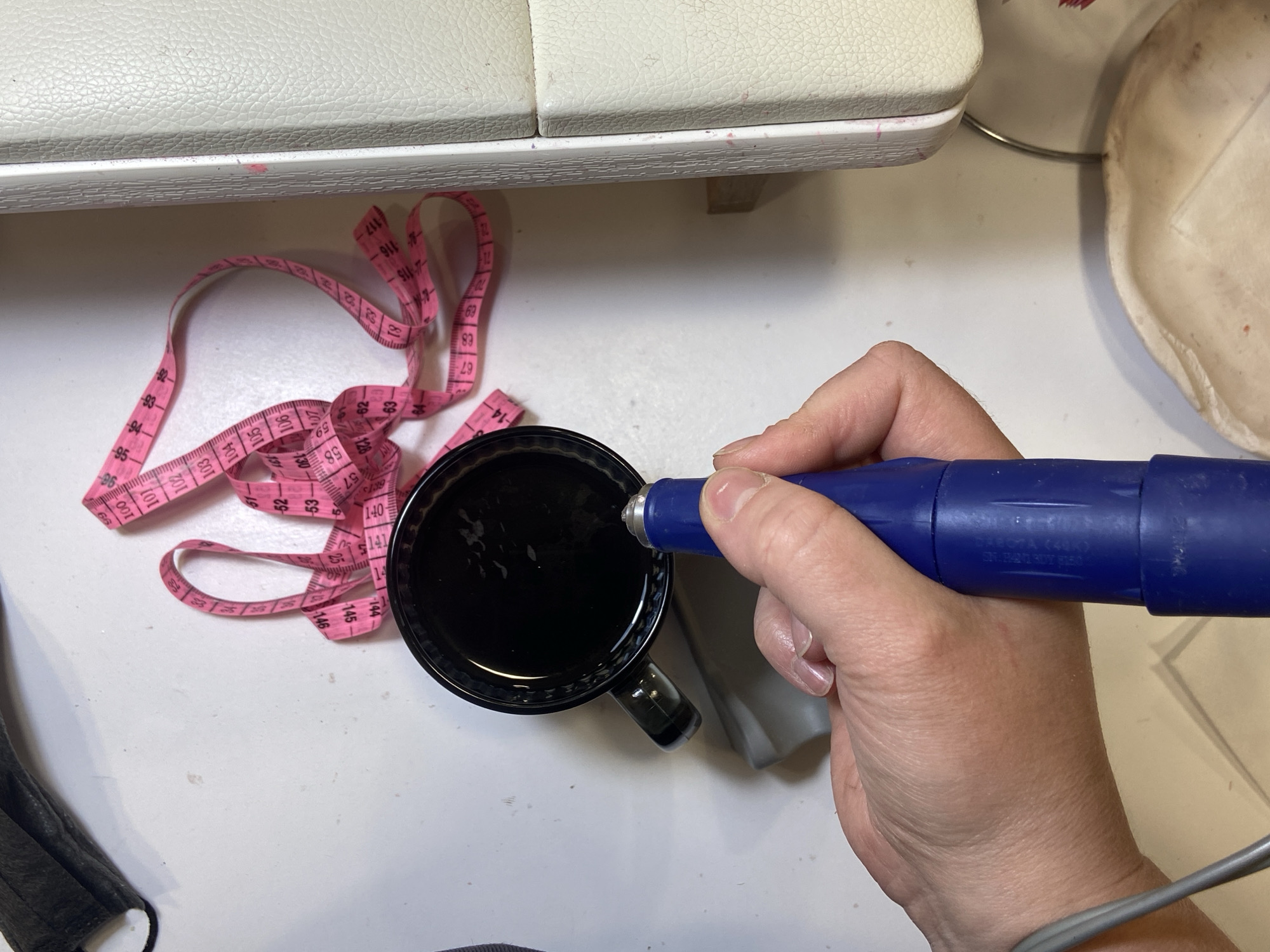 A top-down shot shows a person's hand dipping a blue, cylindrical object, possibly a pen or tool, into a black mug filled with liquid. A pink measuring tape is coiled next to the mug. Part of a sewing machine is visible in the upper left. A gray wire extends from the bottom right. A face mask is partially visible in the bottom left. A porcelain cup is in the top right corner. The scene is set on a white surface. The location in the city of Chaikovsky, Russia is not discernible from the image.