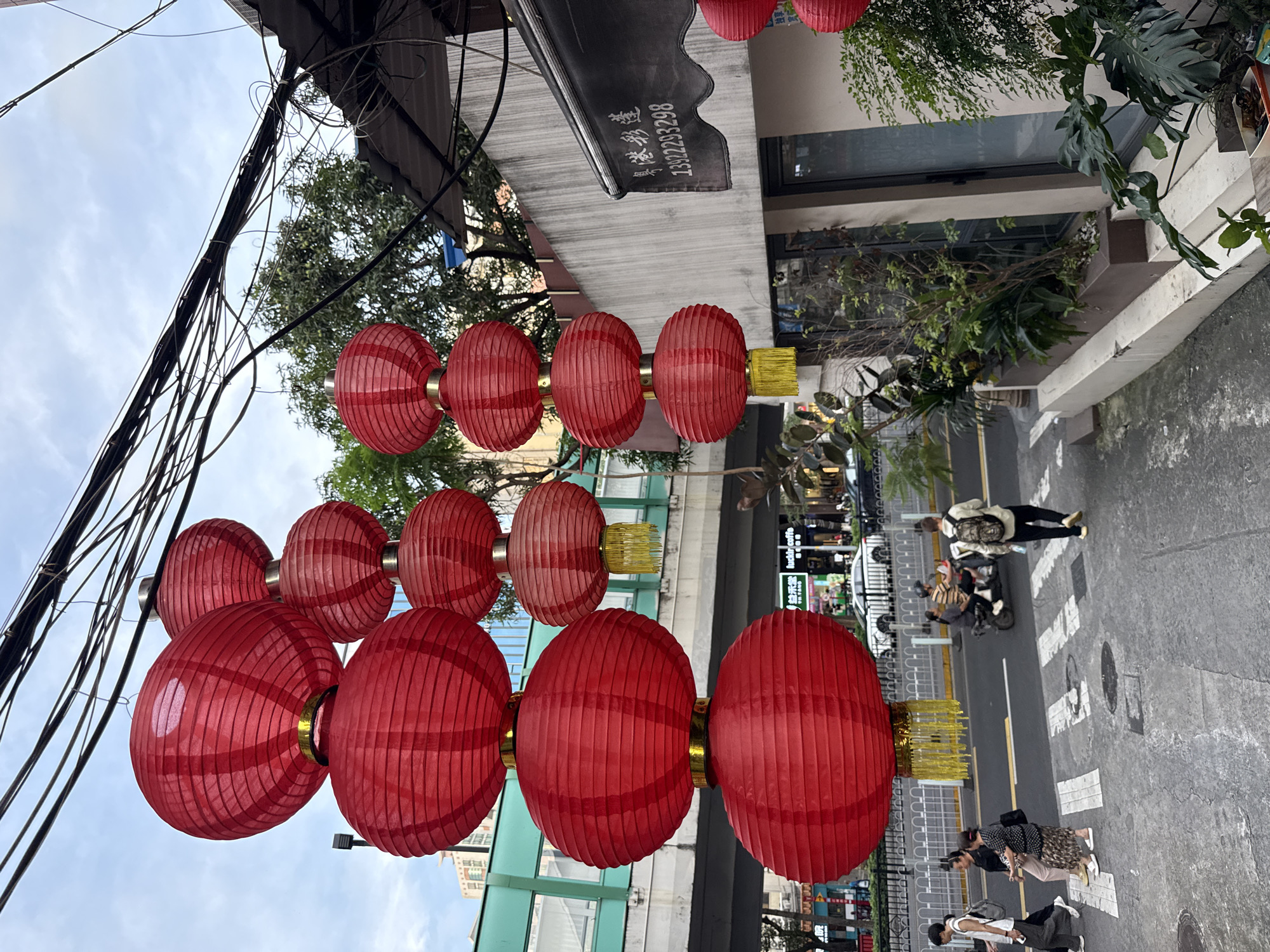 The image shows a street scene in Haizhu District, China, with a focus on hanging red lanterns. Overhead power lines and the awnings of buildings are visible. Below, pedestrians walk on a crosswalk-marked street. Some people are walking individually while others are in small groups. A person on a scooter is also present in the street scene. There are buildings with storefronts and greenery on the sides of the street.