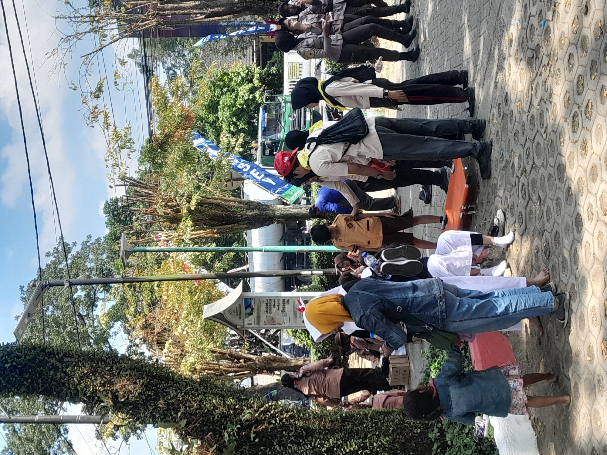 In Genteng Kulon, Indonesia, a group of people, primarily young students in uniform, are gathered outdoors under a sunny sky with scattered clouds. A tall, leafy tree on the left dominates the foreground, with another tree to its right supporting a street light. In the background, a green truck is visible, along with flags and banners. Several students are wearing brown and khaki uniforms, some with yellow scarves, and others are in dark uniforms that appear to be police or military. There's a red stretcher in the middle of the group, suggesting a possible event or activity. A young child in a denim jacket stands in the foreground, looking towards the gathering. The ground is paved with hexagonal tiles.