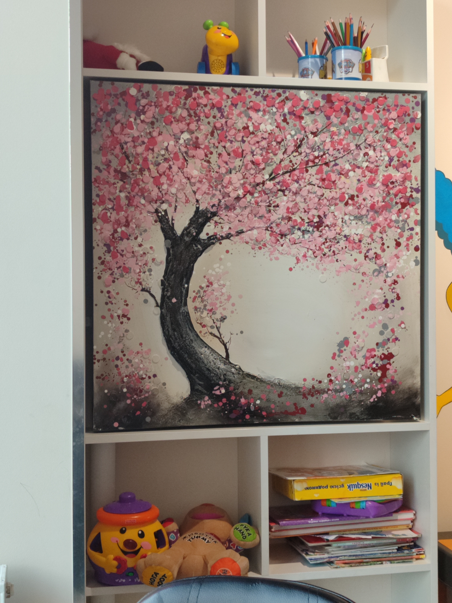 This is a medium shot, eye-level, indoor photograph of a white bookshelf filled with various items. The bookshelf has at least three visible shelves. The top shelf contains a painting of a cherry blossom tree with pink and red blossoms against a grey and white background. To the left of the painting, on the same shelf, is a red and white stuffed object, partially obscured, and a yellow and purple toy with green eyes. To the right of the painting, there are three blue cups filled with colored pencils, and a small white object.
The middle shelf contains a stack of books and a purple toy on top of them. A yellow box with blue text is also on this shelf. To the left of the stack of books, there are two stuffed animals. One is yellow with a purple top and orange trim, and the other is a brown bear-like stuffed animal.
The bottom shelf is mostly obscured by the foreground, but a black chair is visible. The overall environment appears to be a child's room or play area. There are no people visible in the image. The lighting is even and appears to be from an overhead source.