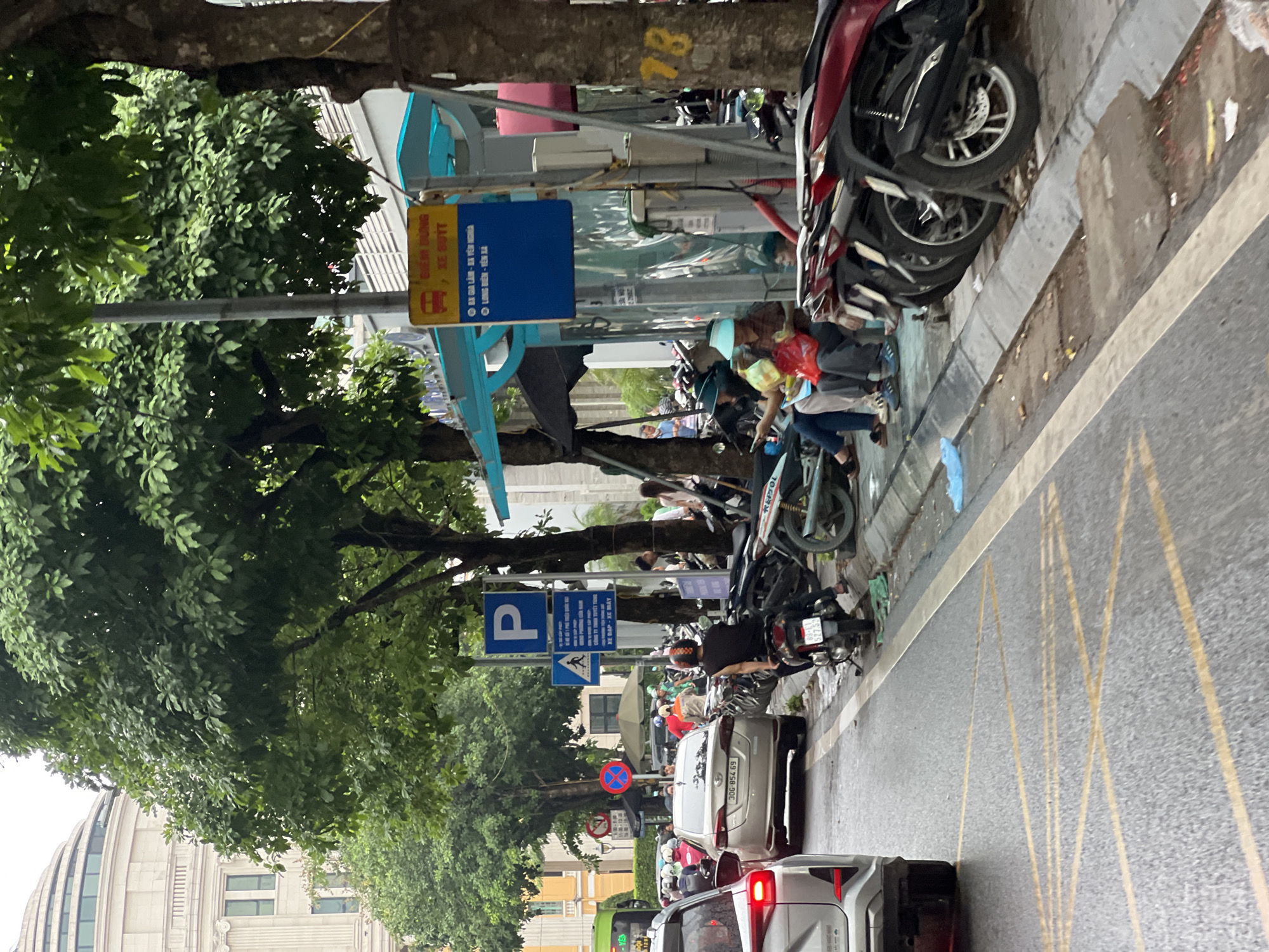 This image depicts a street scene in Hanoi, Vietnam. In the foreground, a silver sedan is stopped in traffic. To the right, a row of parked motorcycles is visible, with several people sitting on the curb next to them. A woman wearing a blue visor and a patterned shirt is holding a plastic bag. A man beside her is wearing a blue shirt and dark pants. Further down the street, more people and vehicles are present.
Several signs are posted. One sign, a blue rectangular sign with white text and a bus icon, indicates a bus stop with route information. Another sign, a blue square with a white 'P', signifies parking. There's also a red circular sign with a diagonal bar, indicating no parking or stopping.
The environment is urban, with trees lining the street and buildings visible in the background. The road appears wet, suggesting it may have recently rained. Yellow lines are painted on the asphalt, indicating traffic lanes and possibly a no-stopping zone. The overall atmosphere is that of a busy street with a mix of vehicles and pedestrians.