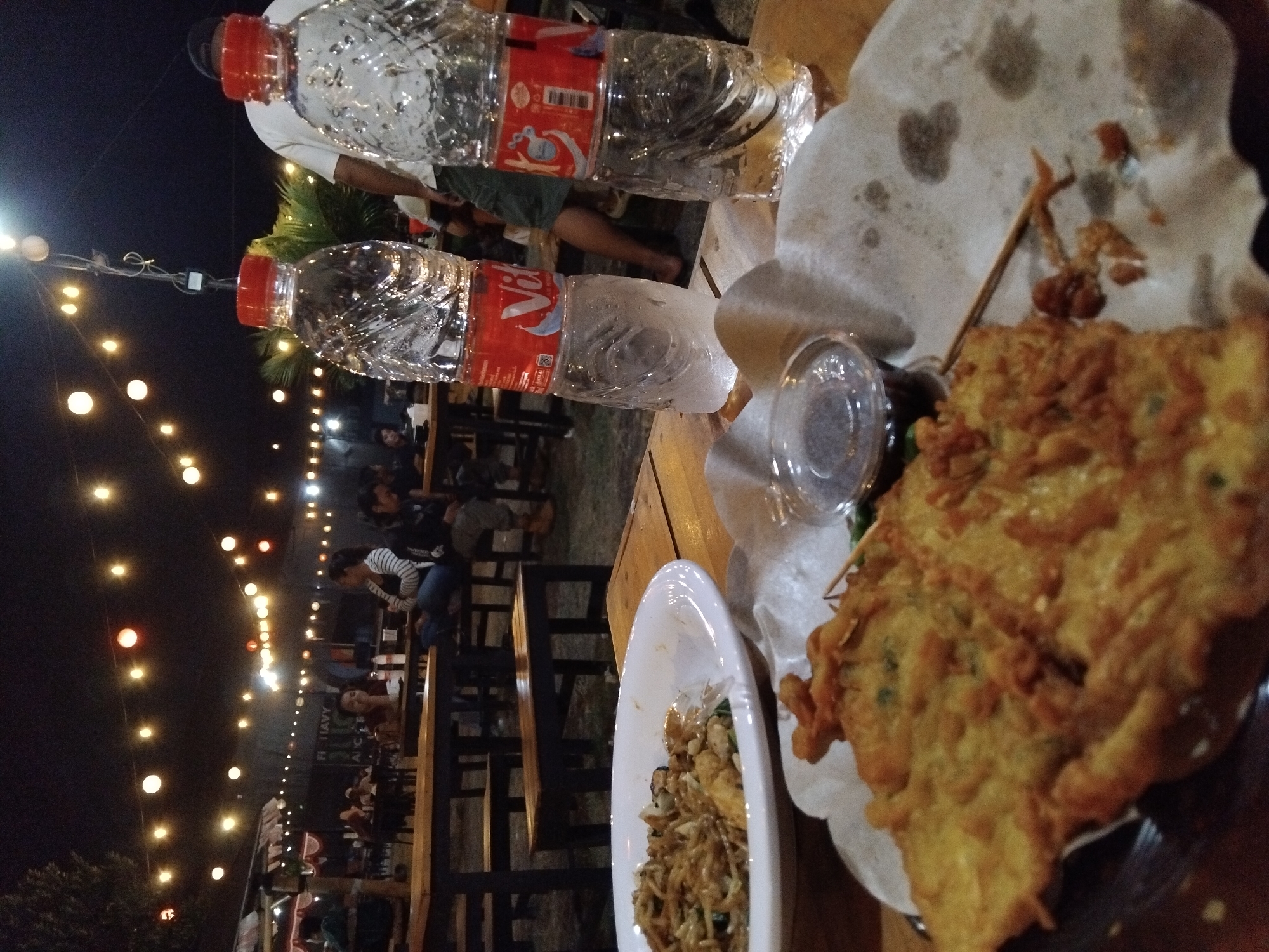 The image depicts an outdoor night scene at a food stall or outdoor dining area. In the foreground, two plastic bottles of water with red caps are placed on a wooden table. One bottle has a label that reads "Vita." Next to the bottles, a plate holds what appears to be fried food, possibly fritters or bakwan, and a small container of dark dipping sauce. Another plate of noodles or fried rice is visible in the background.
The setting is illuminated by strings of warm-toned lights strung across the scene, creating a festive atmosphere. In the middle ground and background, several people are seated at tables, engaged in conversation or eating. Some are silhouetted against the lights. The overall impression is of a casual dining experience in an urban environment at night. The location is specified as the city of Betro, Indonesia.