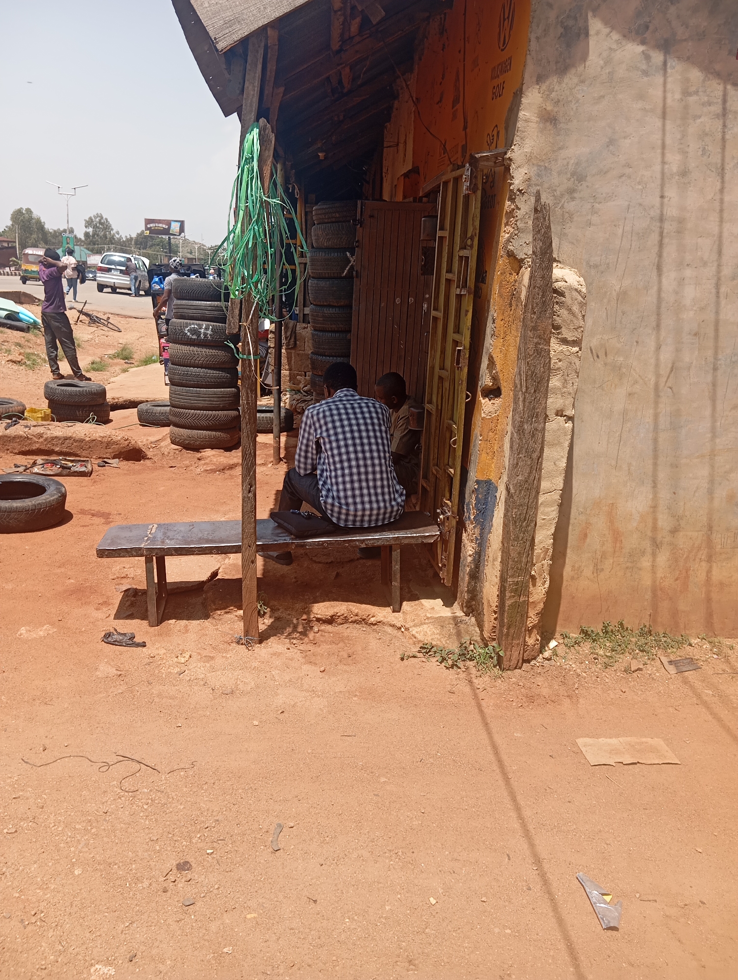 The image depicts an outdoor scene in Jos, Nigeria, likely a tire repair shop or a place where used tires are sold. The setting is rustic, with a makeshift wooden structure providing shade over a workspace. Piles of used tires are stacked high against the wall of the structure, indicating the nature of the business.
In the foreground, a man in a blue and white checkered shirt is seated on a metal bench, facing away from the camera. He appears to be taking a break or waiting. Beside him, a young boy in a brown shirt is also seated, possibly with the man. The ground is reddish-brown dirt, typical of the region, with scattered small stones and some sparse green vegetation.
In the background, a busy street is visible with several vehicles, including a silver car and what appears to be a three-wheeled auto-rickshaw. People are walking around, suggesting it's a commercial area. The sky is bright and appears to be a clear day, suggesting it's daytime. A green plastic ribbon or strap is hanging prominently from the wooden structure.
On the wall of the structure, some faded text is visible. It appears to be a logo with the word "VOLKSWAGEN" and "GOLF" underneath, suggesting that the shop might specialize in tires for these vehicles or is located near a car service center. Another section of text on the wall is less legible but seems to be in a darker color. The overall atmosphere is one of everyday commercial activity in a somewhat unpolished urban environment.