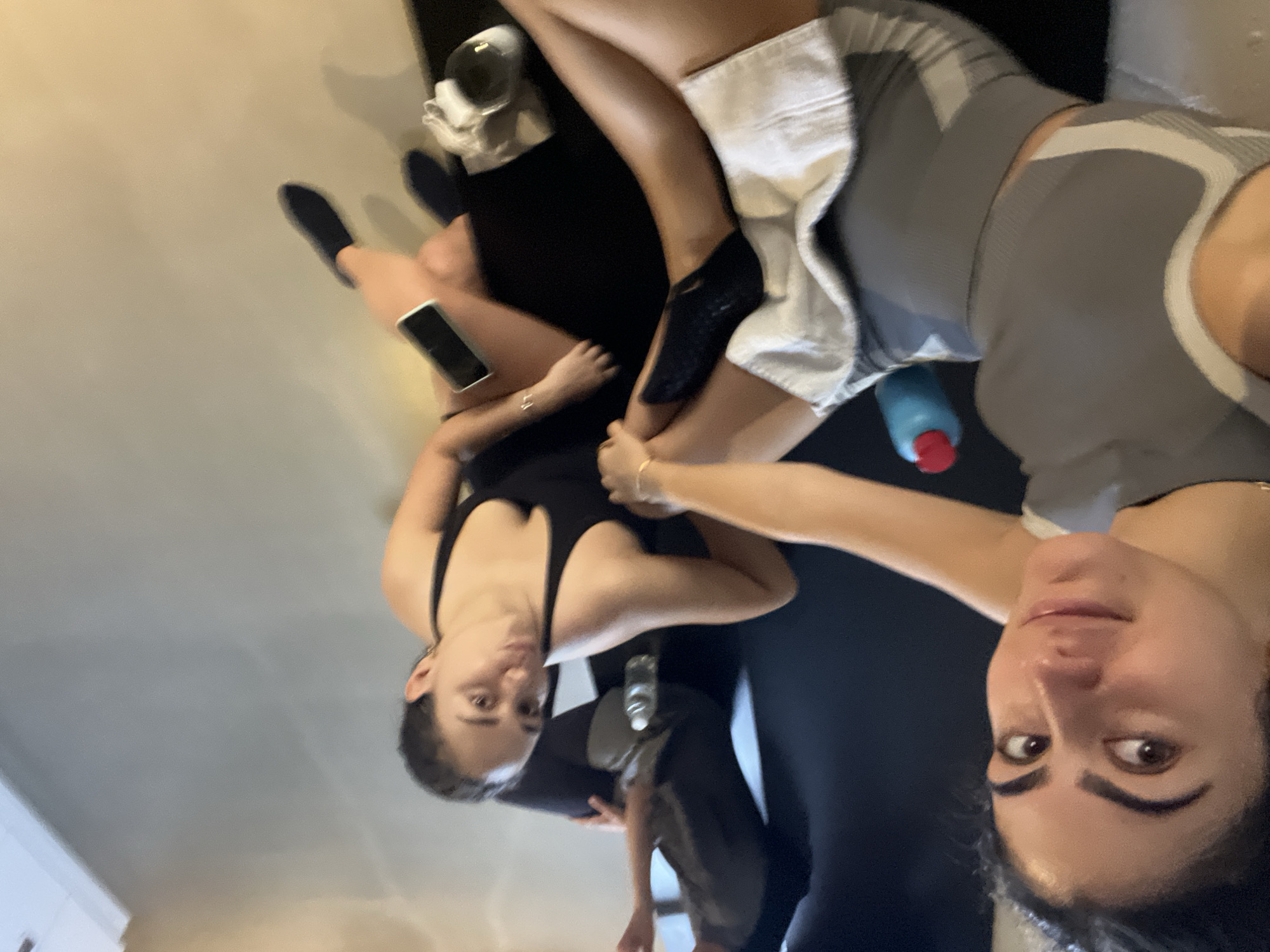 The image shows a group of young women sitting on the floor, presumably in a fitness or yoga studio. The floor appears to be light-colored and smooth, possibly polished concrete or linoleum. In the foreground, one woman is wearing a gray athletic top and shorts, with a white towel draped over her legs. Her legs are extended, and her feet are bare. Next to her, another woman in a black athletic top is sitting with her legs crossed. A third woman, partially visible in the background, is also seated on the floor wearing dark athletic attire. A blue water bottle with a red cap is visible near the woman in the gray top. A smartphone is placed on the floor near the woman in the black top. The overall ambiance suggests a casual, post-workout setting. There is no visual information in the image that can place the scene in Paris, France.