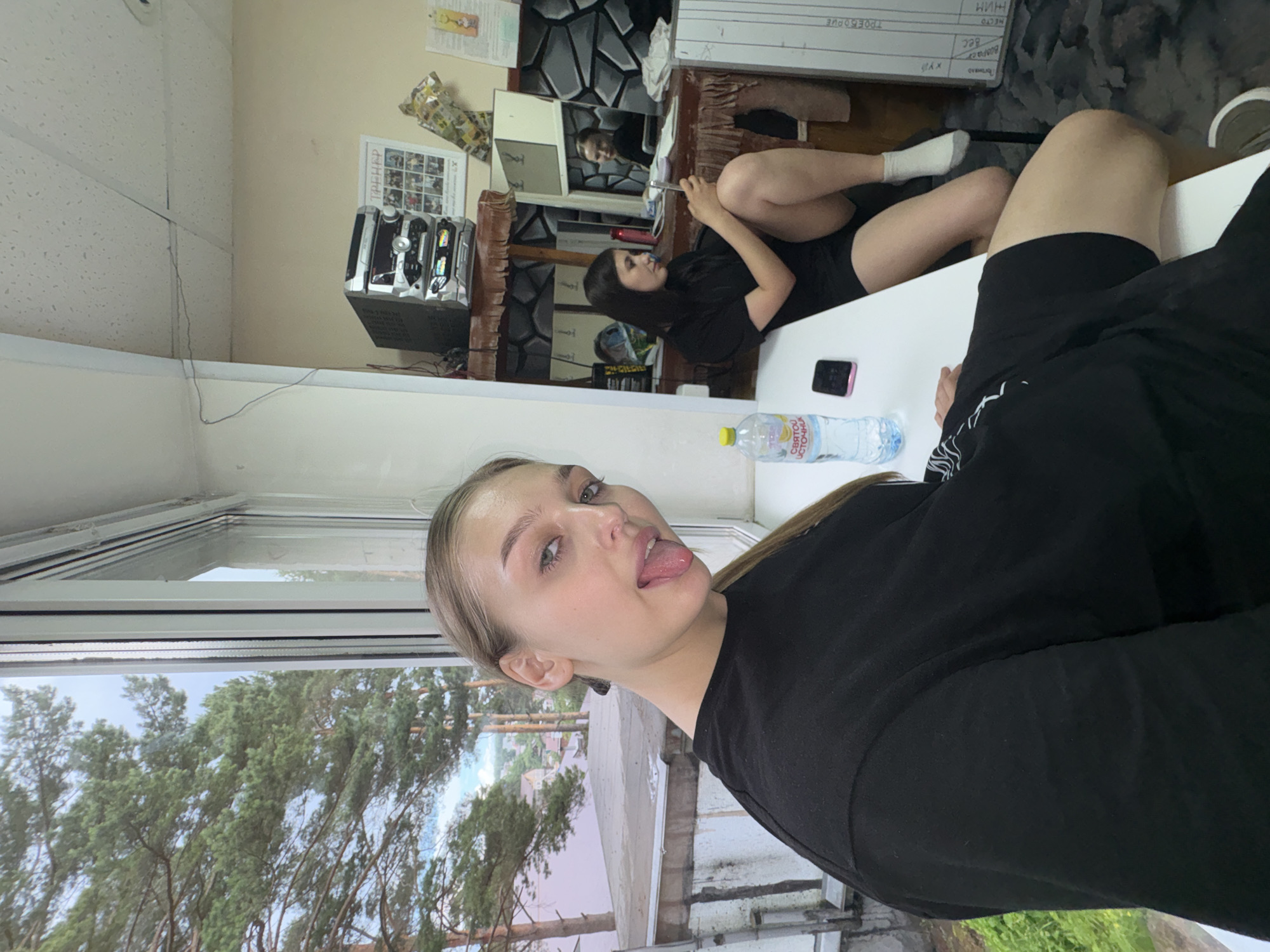In an indoor setting with a window overlooking trees and houses, a young woman with blonde hair pulled back in a ponytail, wearing a black t-shirt, sticks her tongue out at the camera. She is sitting at a white table with a bottle of water and a smartphone. In the background, another young woman with dark hair, also wearing black shorts and a t-shirt, sits with her legs crossed, looking at her phone. A stereo system is visible on a shelf above her. The room appears to be a functional space, possibly an office or a common area.