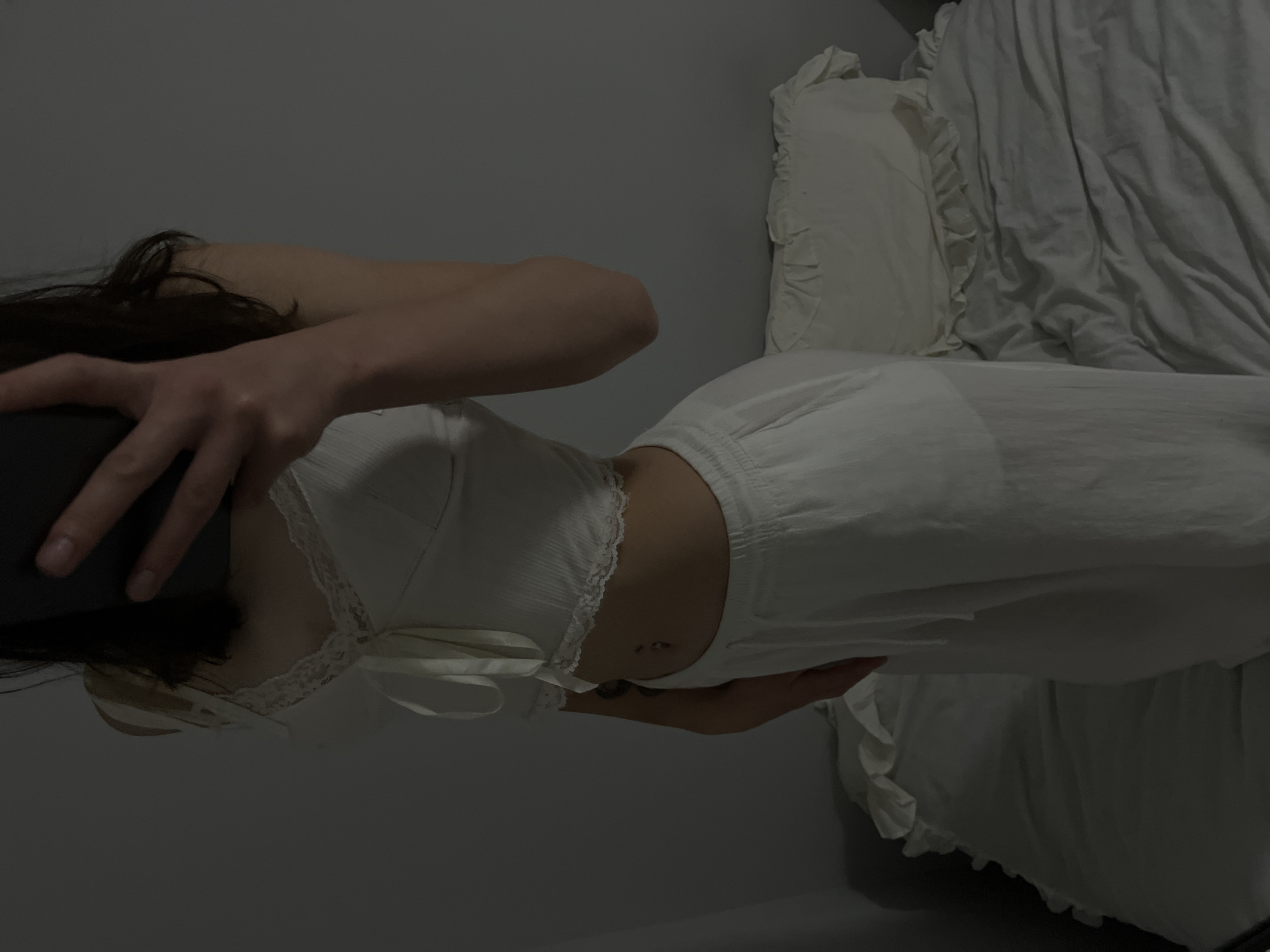 A person, whose face is obscured by a dark object held in their hands, is standing and taking a photo of themselves in what appears to be a bedroom. They are wearing a white lace-trimmed crop top tied at the front and loose white pants. A navel piercing is visible. The person has dark hair and a tattoo on their left arm. They are standing in front of a bed with rumpled white bedding and a pillow with ruffles. The background is a plain, gray wall. The lighting is dim, casting shadows. This image was taken in Ternopil, Ukraine.