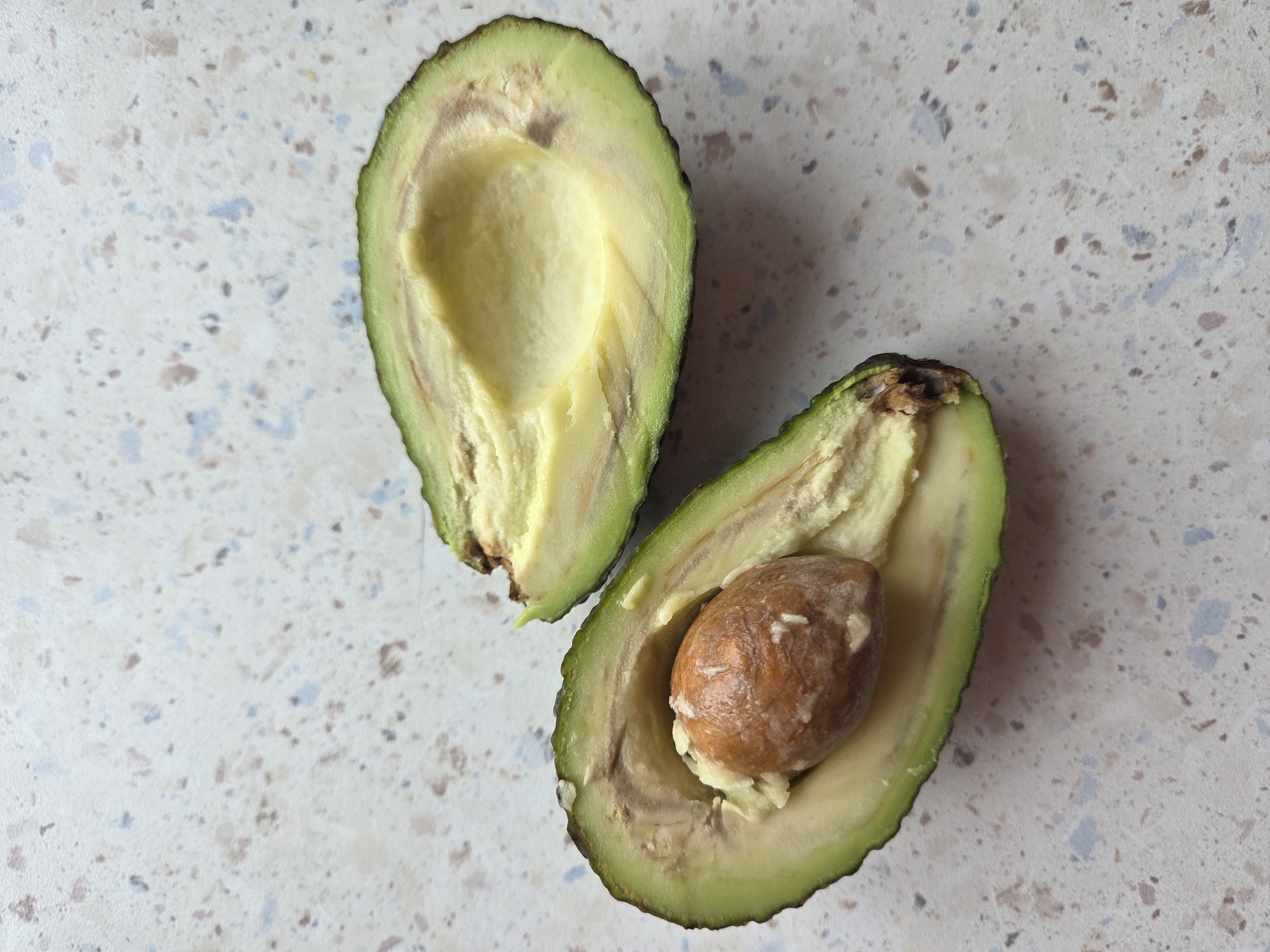 The image shows a halved avocado on a light-colored, speckled surface. One half contains the seed, while the other half is seedless. There are slight discolorations on the avocado flesh. There is no indication that the image was taken in Birobidzhan, Russia.