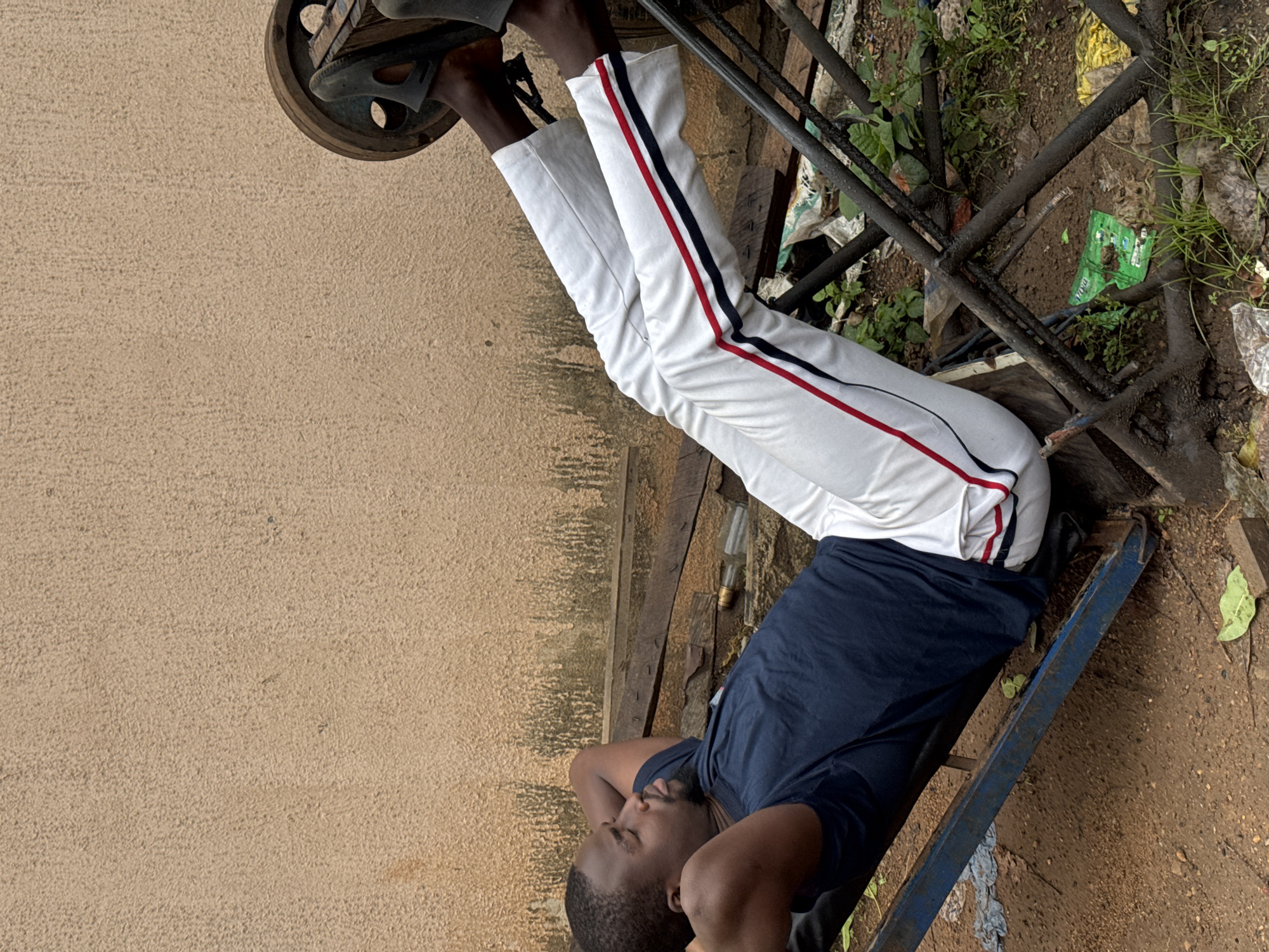 A man is lying down on the ground, appearing to be resting. He is wearing a navy blue t-shirt and white track pants with red and black stripes along the sides. His eyes are closed, and his hands are behind his head, suggesting he is relaxing or possibly sleeping. The man is positioned near a metal structure, which could be part of a bench or some sort of frame.
The ground is dirt and appears to be somewhat uneven, with some patches of grass and fallen leaves. There is also some scattered debris in the background, including plastic bags and what looks like a discarded glass bottle. The overall scene suggests an outdoor setting, possibly in a less-maintained area.
The lighting suggests it could be daytime, but the specific time of day and weather conditions are not discernible from the image. The context of Akwanga, Nigeria, places the scene in a specific geographical location. There is no visible text in the image.