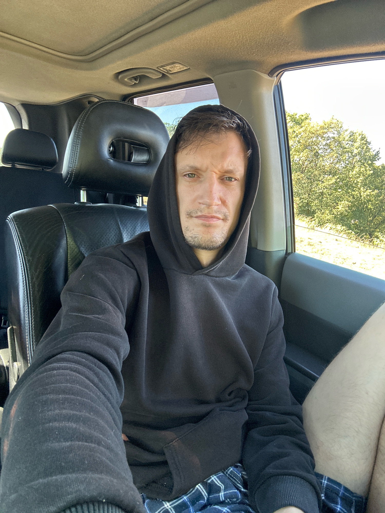 A man with short brown hair and a beard is seated in the passenger seat of a car, looking directly at the camera. He is wearing a black hooded sweatshirt and blue plaid shorts. The interior of the car is visible, including the leather headrest of the seat in front of him, the car's sunroof, and the window to his right, which shows trees and a bright sky outside. The overall scene appears to be a daytime shot taken in a rural area, possibly Makiv, Ukraine.