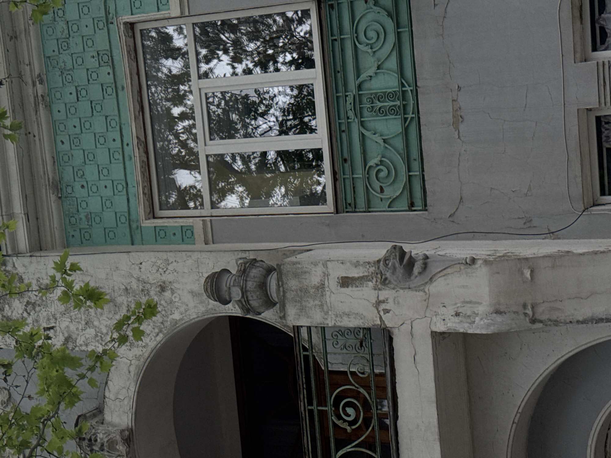 The image shows the exterior architectural details of a building, likely located in Yevpatoriya, Ukraine. The facade features a large window with a white frame, reflecting trees and the sky. A decorative teal-colored grille adorns the side of the window. Below, a white archway is visible, supported by a decorative column and topped with an ornamental figure. The overall structure is weathered with cracked paint and signs of age. Green leaves from a tree appear in the lower-left corner.