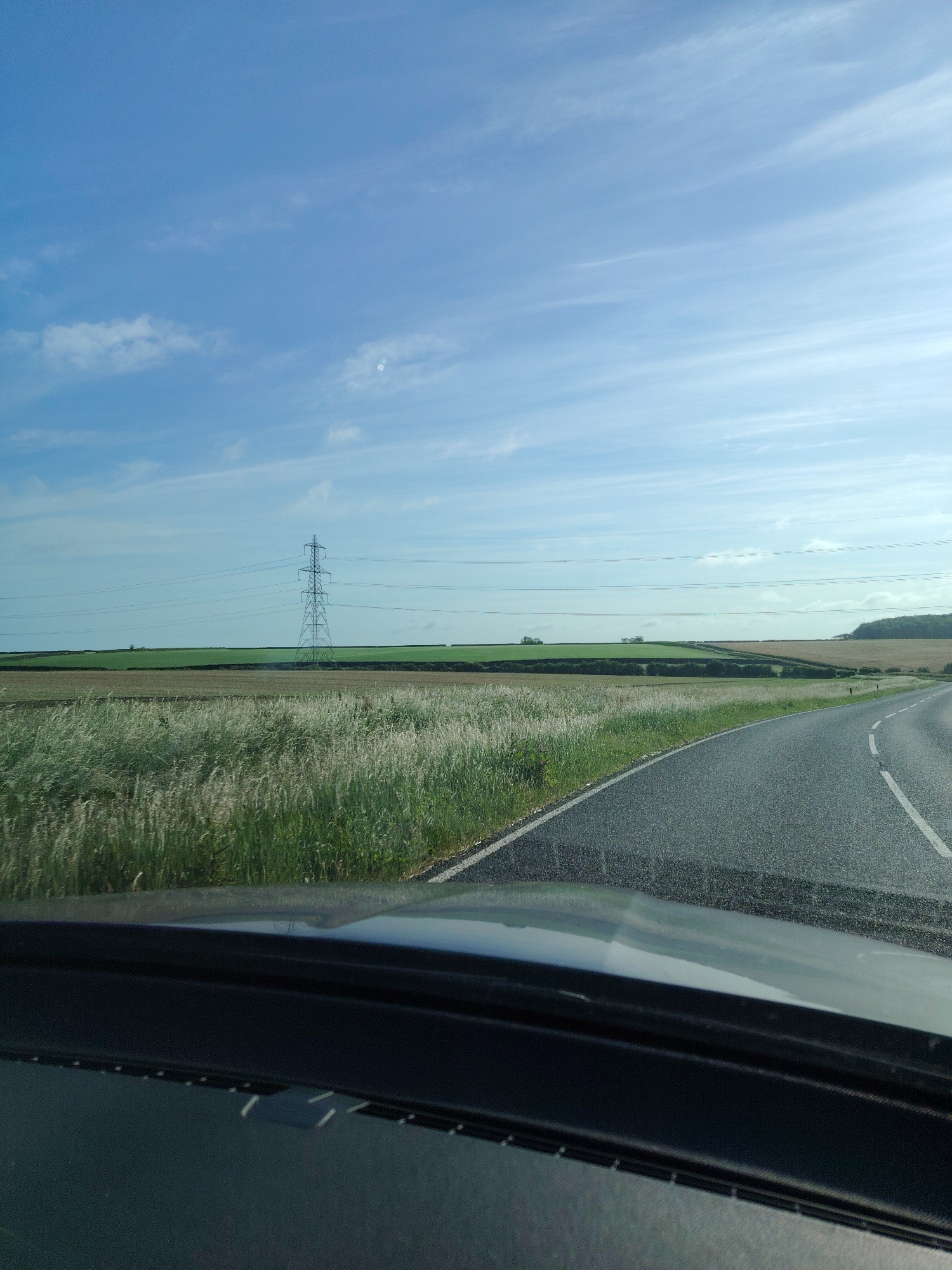 This image captures a rural road scene in Huntingdonshire, United Kingdom, viewed from the interior of a vehicle. The sky, occupying the upper portion, is light blue with scattered white, wispy clouds, and a distinct bright reflection (consistent with lens flare or a reflection of the sun) visible near the top-center-right.
The midground features a gently undulating landscape composed of agricultural fields. A prominent, tall, metal lattice electricity pylon stands in the mid-left section, with multiple parallel power lines extending horizontally across the frame. The fields exhibit varying colors: light green in the immediate left, darker green further back, and a patch of light brown or tilled earth extending towards the right. A distant line of trees and a slightly elevated, densely wooded area are visible on the far right horizon.
The foreground is dominated by a two-lane asphalt road that curves sharply to the right. The road has a solid white line marking its right edge and a dashed white line indicating the center. The left roadside verge is covered in abundant, tall, feathery grasses of light green and beige hues. The bottom section of the image displays the dark grey dashboard and the lower part of the windshield of the vehicle, showing a dark trim piece and faint reflections on the glass. No individuals or discernible activities are present in the scene.