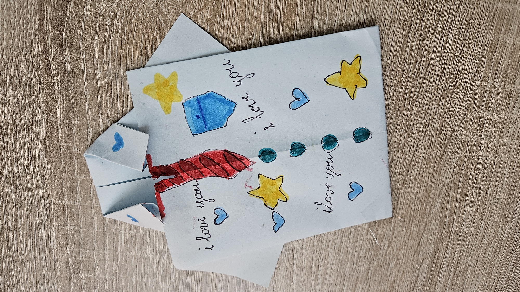A handmade origami paper shirt with a red tie is decorated with hand-drawn elements on a wooden surface. The shirt is light blue and folded to resemble a collared shirt. A red tie with black stripes is drawn down the center. The front of the shirt is decorated with blue hearts, yellow stars, and teal circles. The phrases "i love you" are written in black cursive script multiple times on the shirt. A blue pocket with a black outline is drawn on the right side of the shirt. The wooden surface appears to be a tabletop with a visible wood grain pattern. No people or other objects are visible in the image. The image does not provide any information to place it in Bratislava, Slovakia.