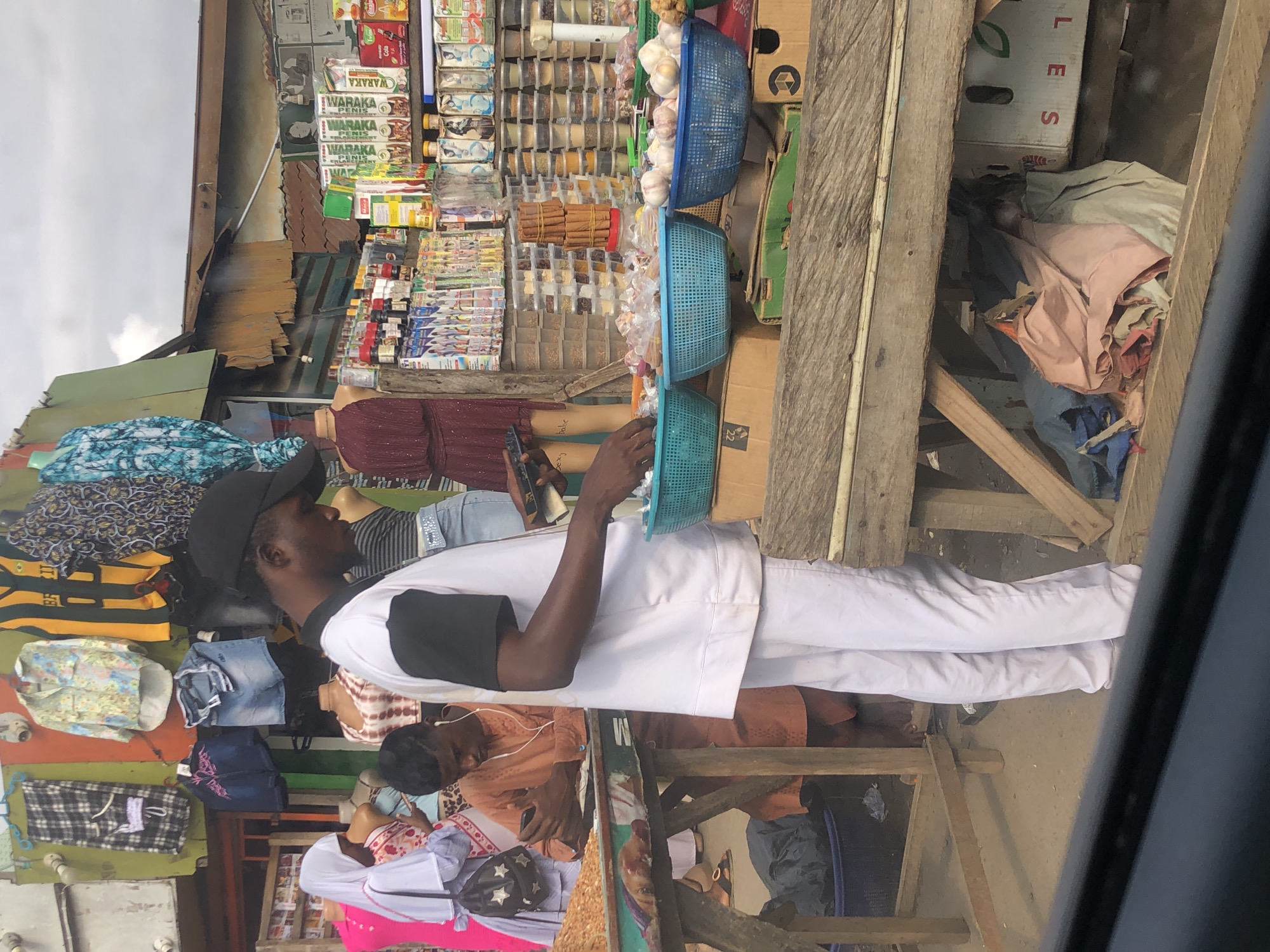 In Lagos, Nigeria, a market scene unfolds under an overcast sky. A young man, dressed in white trousers and a white lab coat with black sleeves, stands in the foreground, his gaze fixed on a mobile device in his hand. Beside him, another young man in an orange outfit, wearing earbuds, is also engrossed in his phone. They are positioned behind a stall displaying a variety of goods.
The stall is laden with fresh produce, including baskets of garlic and what appears to be nuts or seeds. Behind the produce, shelves are stocked with numerous small containers, likely holding spices or other commodities. Above these, rows of packaged goods, including what looks like medicinal or health products and beverages, are neatly arranged. Further up, clothing items, such as patterned shirts and denim shorts, are displayed on hangers, suggesting a diverse market offering.
The market structure itself is rustic, with corrugated metal roofing and wooden stalls, indicative of a bustling, open-air marketplace typical of Lagos. The ground is paved with concrete. In the background, other market-goers are visible, including a woman in a white hijab. The overall impression is one of everyday commerce in a vibrant urban setting.