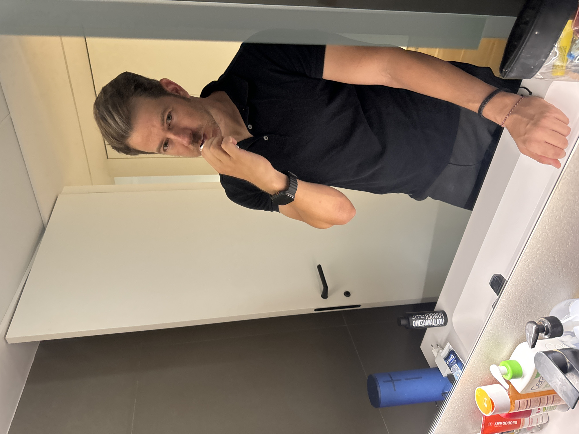 A man with short, styled blonde hair and blue eyes is brushing his teeth in a modern bathroom. He is wearing a black polo shirt and dark pants. He has a watch on his left wrist and two bracelets on his right wrist. The bathroom features a white countertop with a sink, a mirror, and various toiletries including a blue speaker, toothpastes, and soap dispensers. A white door with a black handle is visible in the background. The scene appears to be indoors, likely a personal residence or hotel. There is no information to suggest this location is in Murcia, Spain.