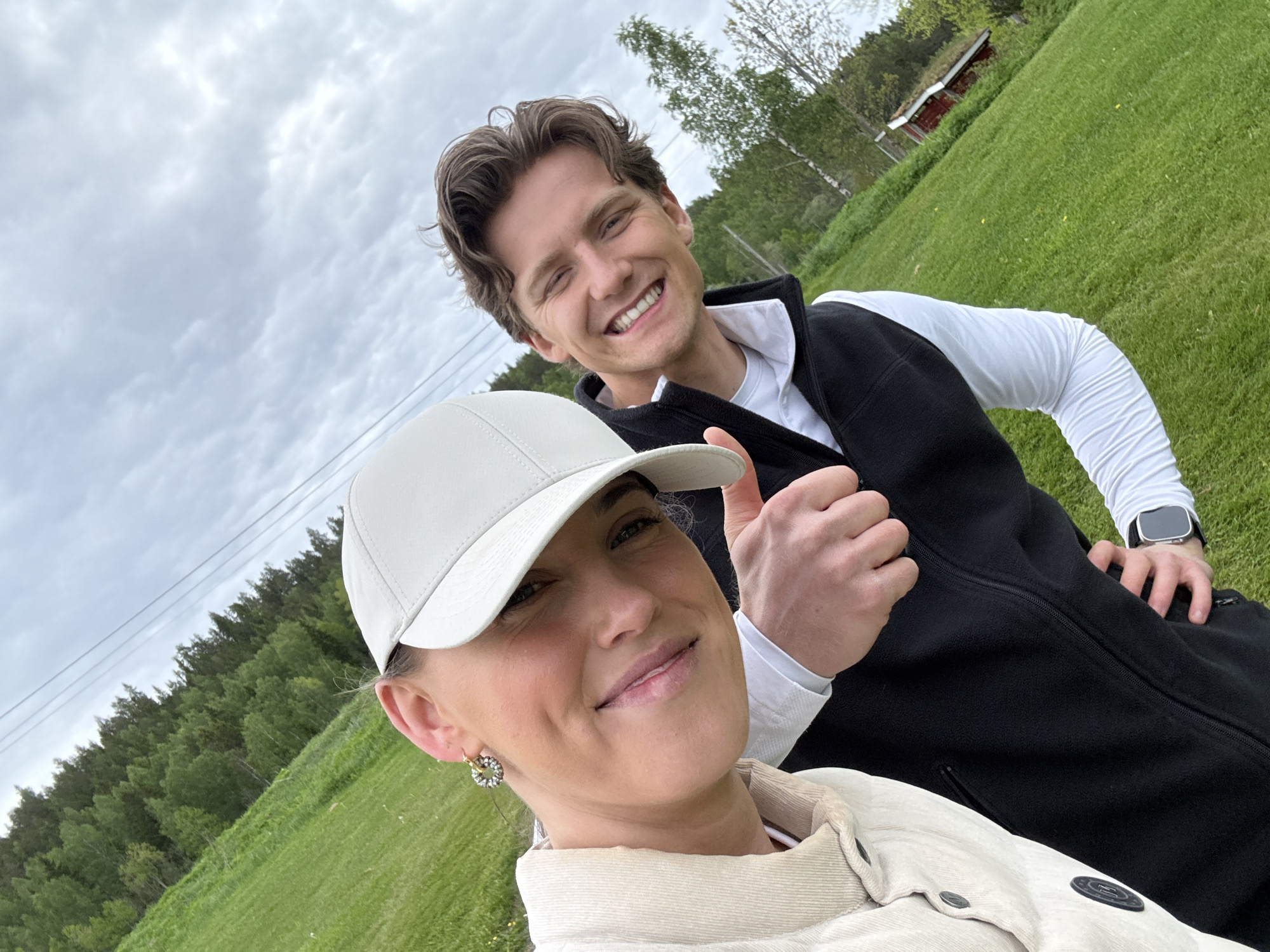 A selfie taken in a grassy area in Løvstad, Norway, shows two people. A woman in the foreground is wearing a light-colored jacket and a white baseball cap. She has an earring in her left ear and a smile on her face. Behind her is a man in a black vest over a white long-sleeved shirt, giving a thumbs-up. He is also smiling. The background includes a grassy field, trees, and a small red building with a sod roof in the distance. The sky is overcast.
