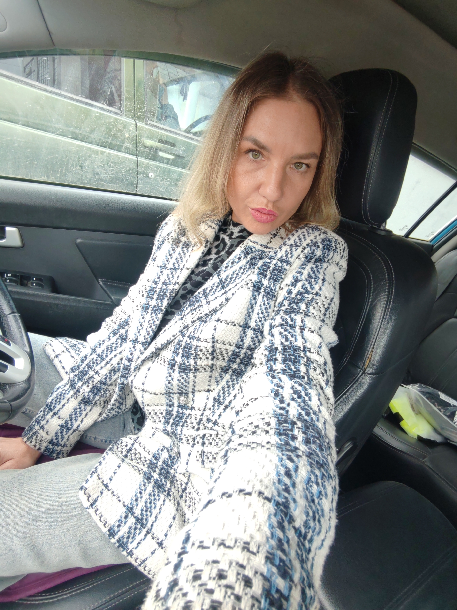 A blonde woman is sitting in the driver's seat of a car in Petropavlovsk-Kamchatsky, Russia. She is wearing a black and white plaid jacket over a leopard-print top and light-wash jeans. The car's interior is visible, including the steering wheel, dashboard, and black leather seats. The window on the driver's side shows a view of other vehicles and buildings, suggesting an urban environment. The woman is looking at the camera and appears to be taking a selfie. The lighting suggests it is daytime, possibly overcast.