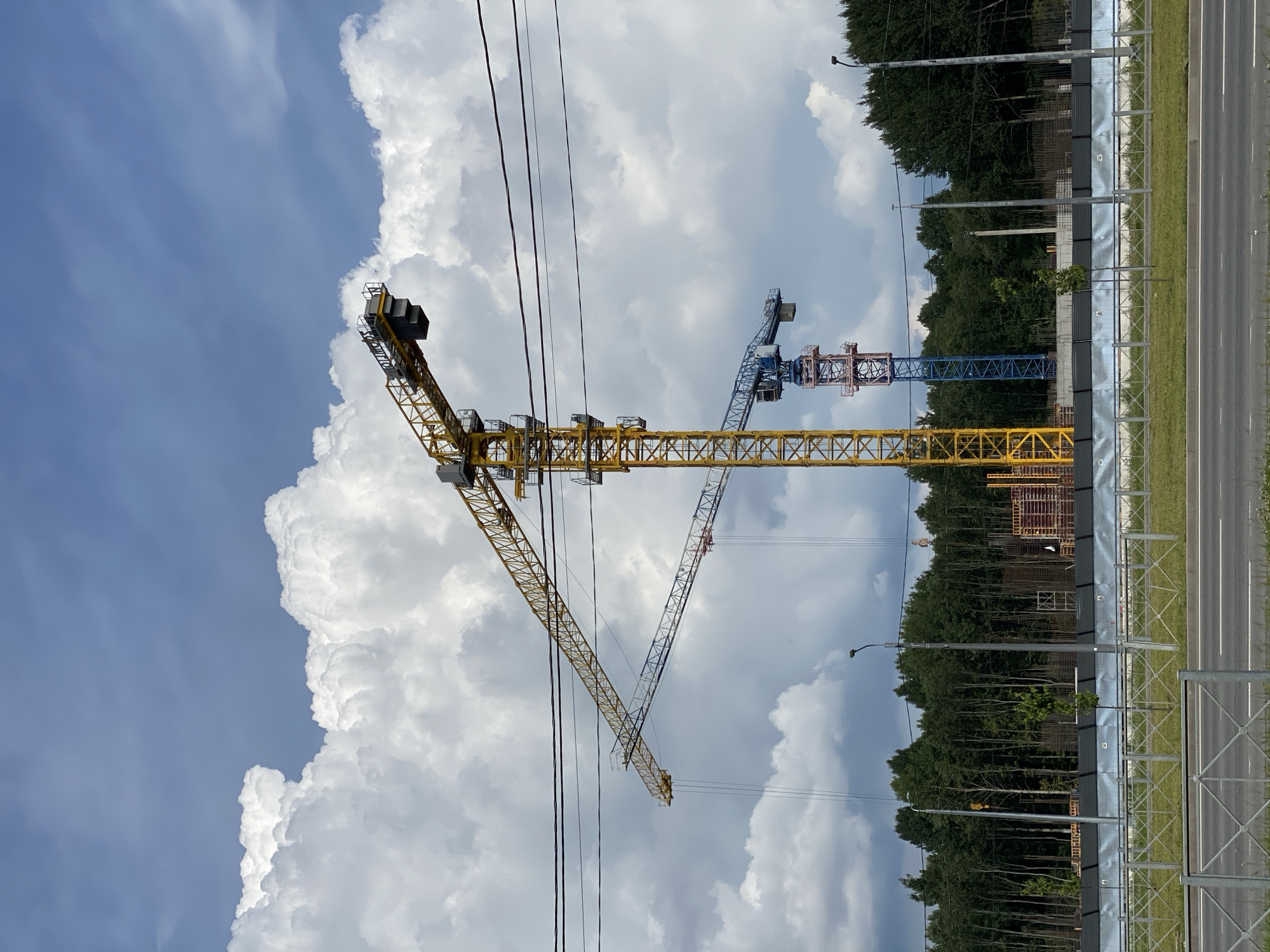 A daytime, eye-level, medium shot captures a construction site under a cloudy blue sky in Cherepovets, Russia. In the foreground, a paved road runs horizontally, bordered by a grassy median and a metal fence. Behind the fence, a construction barrier is visible, with a dense line of trees forming the backdrop. Dominating the scene are three tower cranes. The largest, a bright yellow crane, stands prominently in the center, its arm extended across the frame. To its left, a second, smaller crane, primarily blue with red accents, is positioned further back. A third crane, mostly obscured by the other two and appearing smaller and lighter in color, is visible to the far left. The sky is filled with large, fluffy white clouds against a bright blue expanse. Power lines are strung across the upper portion of the image.