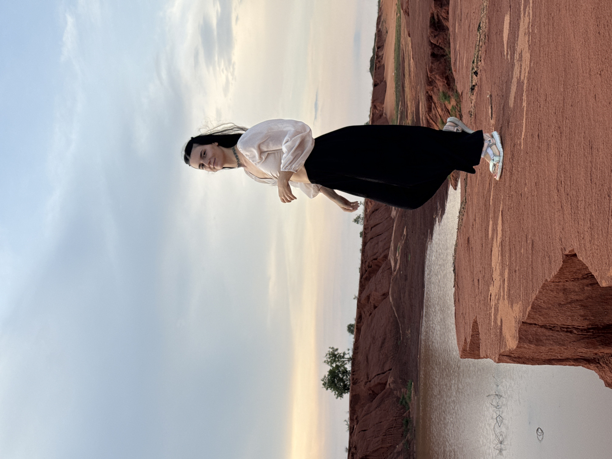 A woman stands posing amidst the distinctive red rock formations and water of what appears to be the Fairy Stream in Mui Ne, Vietnam, under a soft, overcast sky suggesting late afternoon or early evening. The image is oriented vertically with the horizon line running through the middle.
The main subject is a young woman with dark, shoulder-length hair parted down the middle, which appears to be slightly wind-blown. She is wearing a light, long-sleeved, off-white blouse with a V-neck, tucked into a flowing black skirt or wide-leg trousers. A silver-colored chain necklace is visible around her neck. She has a gentle smile and her gaze is directed slightly to her left, with her body angled towards the right. Her right hand is slightly raised, and her left arm is partially obscured by her body. She wears light-colored, possibly white or beige, strapped sandals on her feet.
She is positioned on the edge of a vibrant reddish-brown sand and rock bank, which dominates the lower right portion of the image. The texture of the rock appears weathered and layered. Below this bank, to her right, is a body of calm water, reflecting the soft light from the sky. On the opposite side of the water, more reddish rock formations are visible, with sparse green vegetation, including a few small trees, growing along the banks and on the distant ridges. The sky occupies the upper left portion of the image, displaying soft grey and pale yellow hues, indicating a cloudy yet bright atmosphere. There is no visible text in the image.