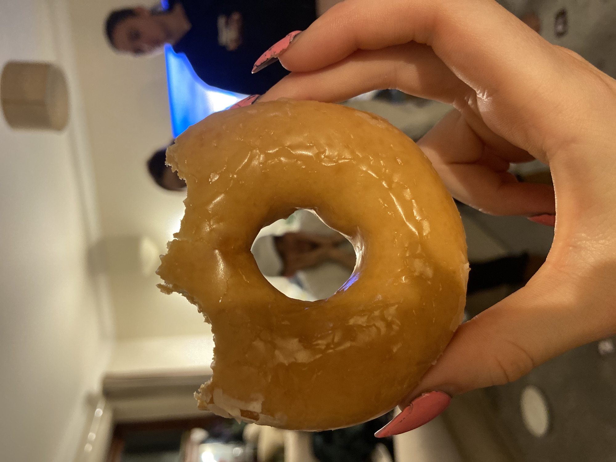 A partially eaten glazed donut is held in a person's hand with pink nail polish. In the background, two people can be seen in a room with a television. A lamp is visible on the wall. The location of Reigate and Banstead, United Kingdom is not discernable from the image.