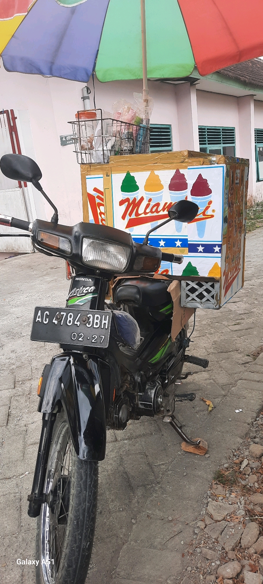 A motorcycle is parked on a paved surface in front of a light pink building with windows. The motorcycle is black and has a license plate that reads "AG 4784 3BH" and "02-27". A large, colorful umbrella is attached to the back of the motorcycle, providing shade over a box adorned with images of ice cream cones. The box appears to be a mobile ice cream vending unit. There are various items in a basket attached to the front of the ice cream unit, including plastic bags and containers. The environment suggests an outdoor setting in a residential or commercial area. The image is a full-frame photograph taken from a low angle.