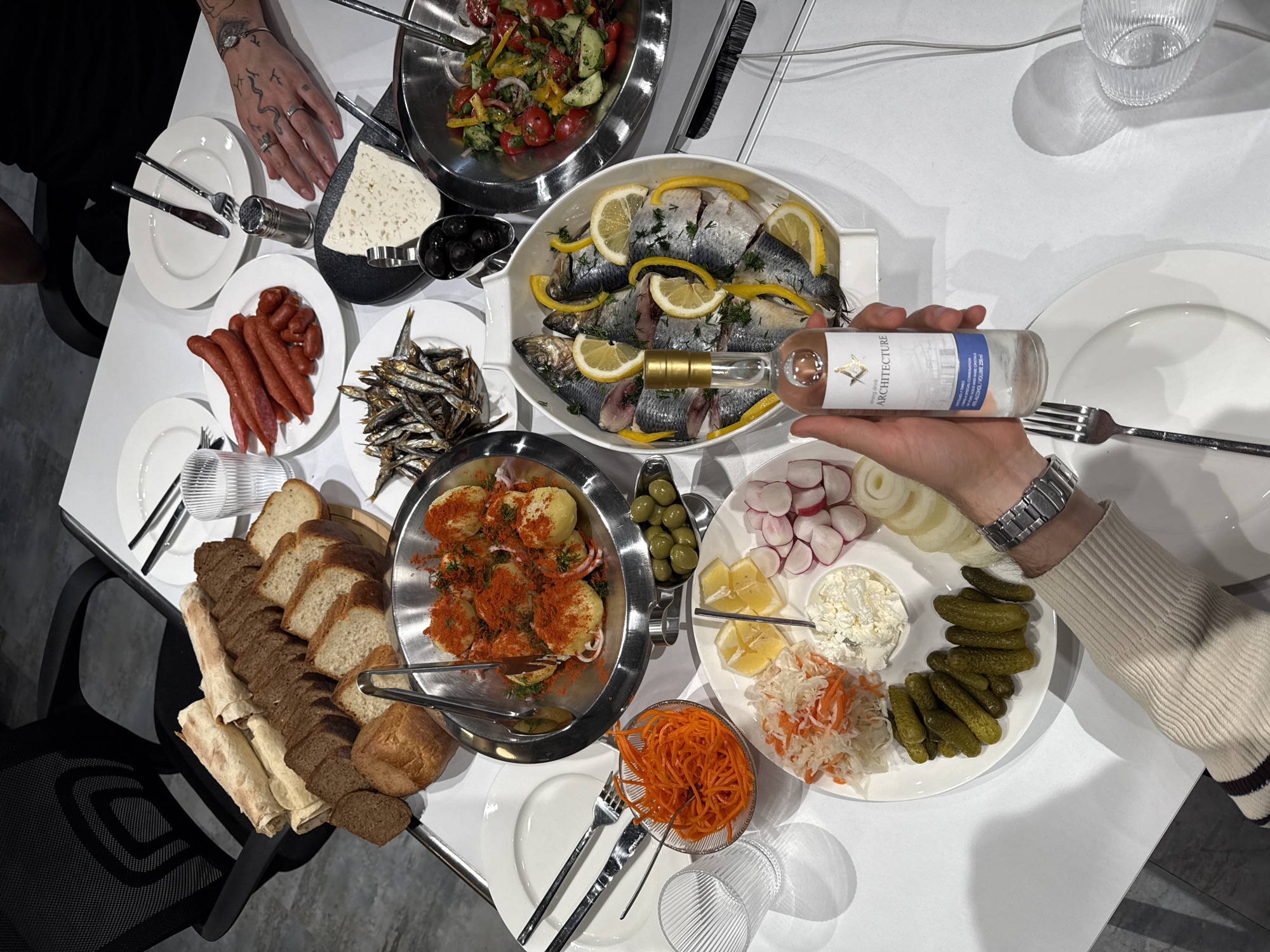 The image shows a table laden with food, presumably in a home in Yerevan, Armenia. The table, covered with a white tablecloth, is crowded with various dishes. Visible are plates with fish, sausages, sliced bread, potatoes, carrots, pickles, radishes, onions, salads, and a bowl of olives. Several people are present, including at least one person with tattoos on their hands. Someone is holding a bottle of "Architecture" wine near a plate of fish. There are plates and silverware arranged for dining. Glasses sit on the table. The setting is indoors, and the floor is a light gray color.