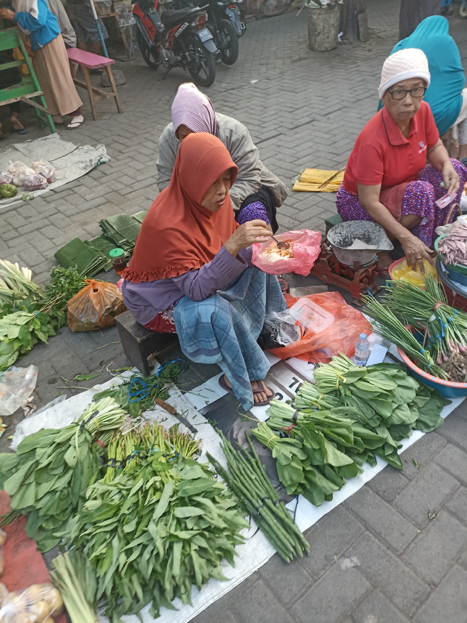 In Kelurahan Kepatihan, Indonesia, three women are engaged in commerce at an outdoor market. Two women are seated on the ground, surrounded by fresh produce. One, wearing an orange headscarf and a purple shirt, is actively preparing food from a pink plastic bag. The other, to her right and wearing a white knitted cap and glasses, is seated with a red shirt and a patterned skirt, looking towards the viewer. A third woman, partially visible in a teal headscarf, is seated further back. The ground is paved with gray bricks. Piles of green vegetables, including leafy greens and what appear to be bundles of spring onions, are neatly arranged on white cloths and in plastic bags. Several motorcycles are parked in the background, suggesting a busy market setting. The overall scene depicts a typical day at a local Indonesian market.