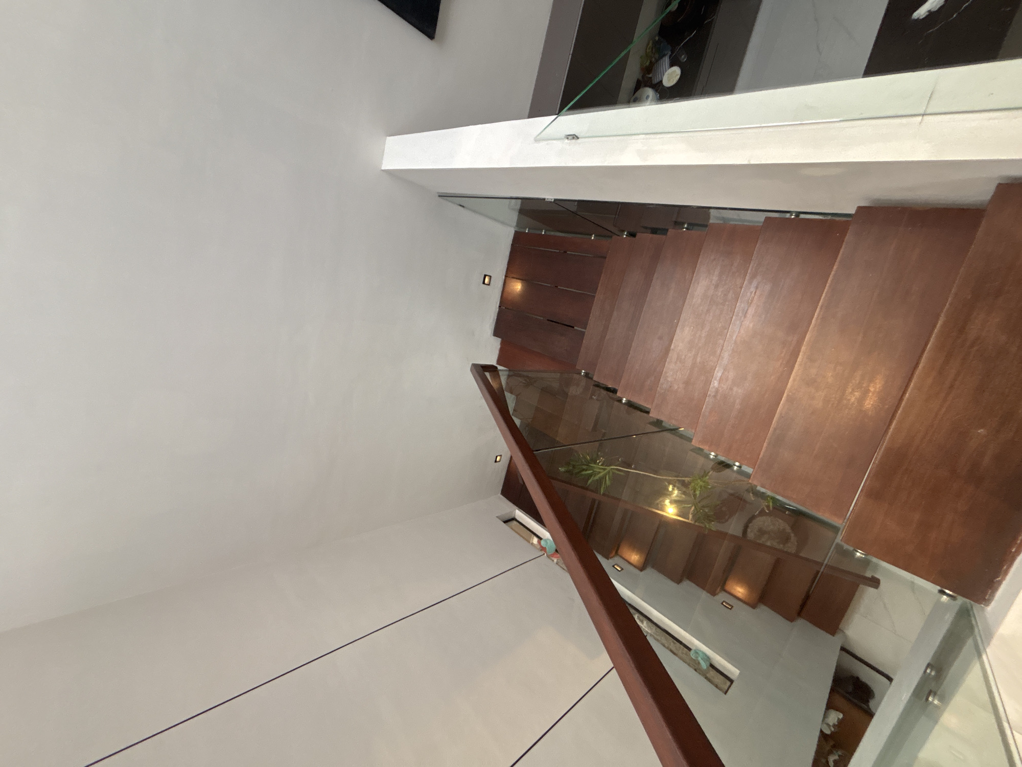 The image shows a modern interior staircase with wooden steps and a glass railing. The staircase is located in a home in North Kuta, Indonesia. The walls are painted white, and there is a wooden handrail on one side of the stairs. A small plant is visible on one of the lower steps. The flooring on the lower level appears to be tiled. The overall aesthetic is minimalist and contemporary.