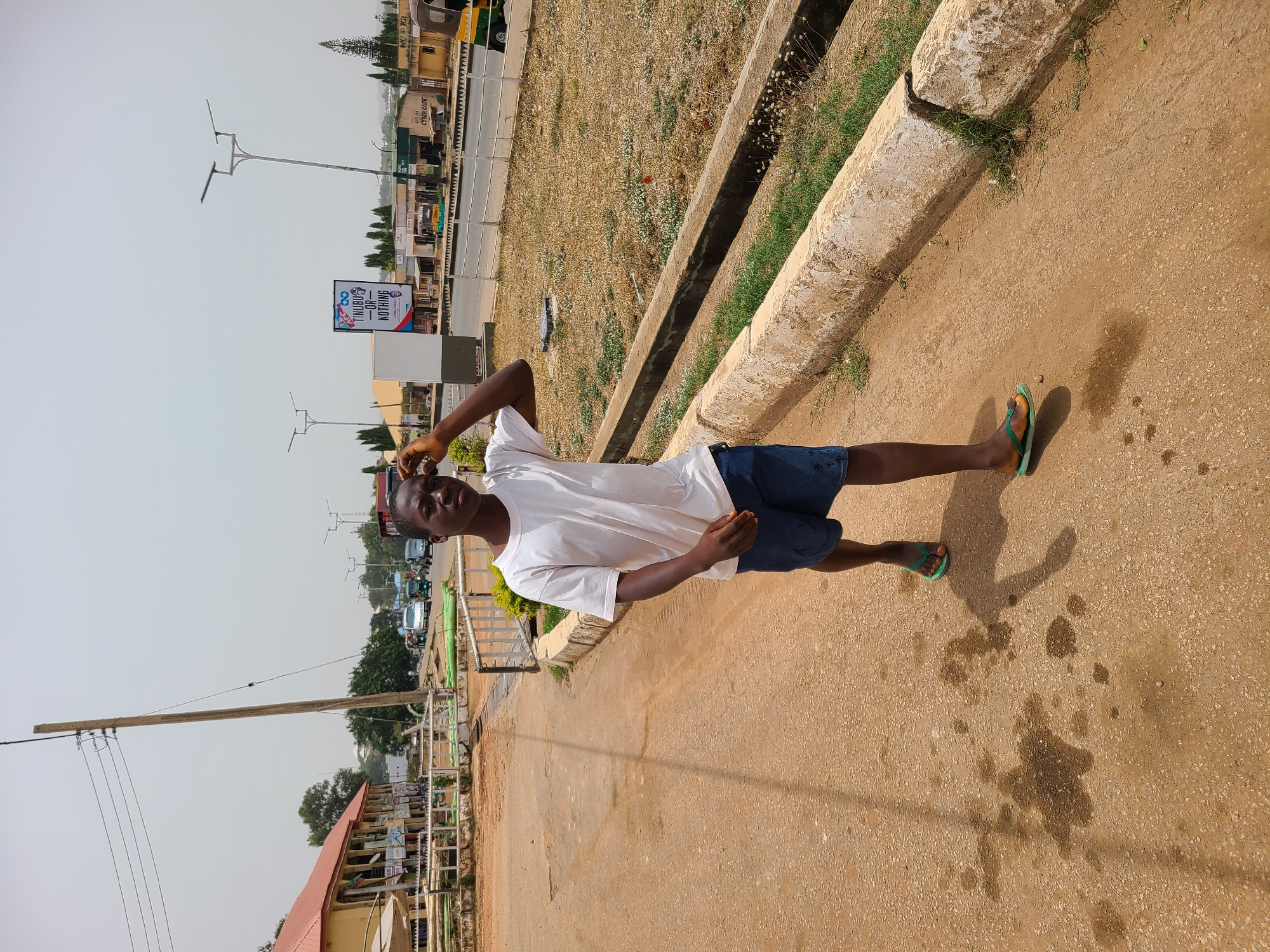 This outdoor image, likely taken in Jos, Nigeria, features a young Black male standing on a sunny day. He is positioned slightly to the left of the frame's center, looking towards the camera with a relaxed expression. He wears a white t-shirt, dark blue shorts, and green flip-flops, with one hand casually raised towards his head.
The setting appears to be a semi-urban environment. The man stands on a wide, light brown, unpaved or lightly paved path. To his right, a concrete curb or drainage ditch runs parallel to the path, separating it from an area of dry, yellowish grass and sparse weeds. Beyond this, a paved road is visible with several vehicles, including cars and tricycles. A row of buildings of varying sizes and designs lines the far side of this road.
The sky is bright and clear, indicating full daylight, likely mid-morning or early afternoon given the distinct shadows cast on the ground by the person and other objects. Several wooden utility poles with electrical wires are visible against the sky, adding to the urban landscape.
A prominent detail in the background is a billboard displaying the text "TINUBU OR NOTHING," likely a political slogan. Below this, another sign partially reads "CYBER CAFE." The overall atmosphere is bright and warm, capturing a typical daytime street scene.