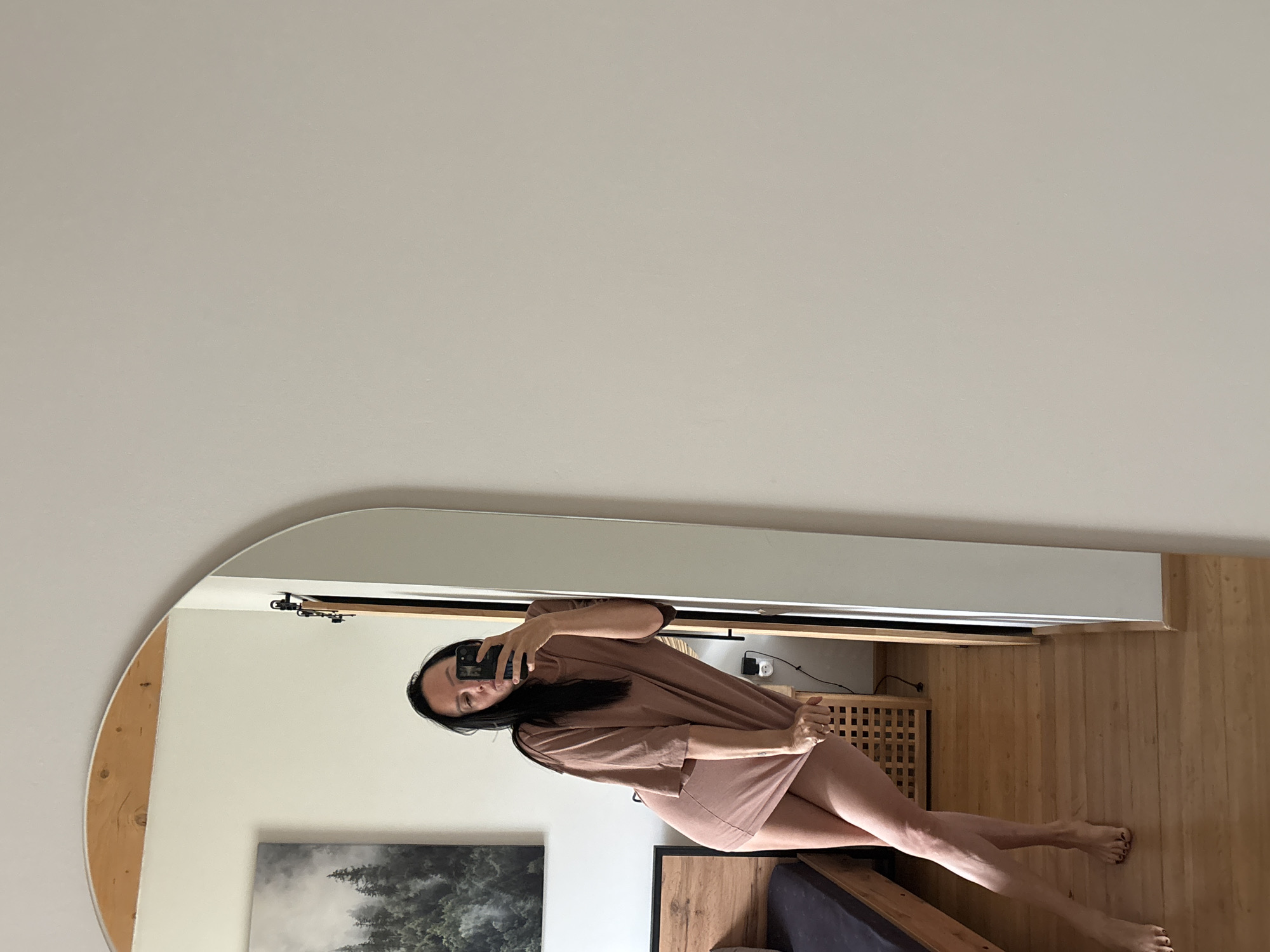 A woman is taking a mirror selfie in a bedroom. She is leaning against a large, arched mirror, with her body angled towards the camera. She is wearing a loose-fitting, light brown t-shirt dress that exposes her legs. Her right hand is lifting the hem of the dress. She has long, dark hair and is holding a black smartphone in her left hand, using it to take the photo.
The bedroom appears to be furnished with a wooden bed frame on the left side of the image. A dark colored mattress is on the bed. Behind the woman, a wooden wardrobe or cabinet with a slatted front is visible. A framed picture of a forest landscape hangs on the wall above the bed. The room has wooden flooring and white walls. The mirror has a wooden frame at the top.
The image is a reflection in a mirror, showing the woman and part of the room. The right side of the mirror shows a plain white wall, suggesting the room is not fully captured or the wall is a prominent feature. The overall lighting is bright and natural. There is no indication of the city of Chaikovsky, Russia in the image itself.