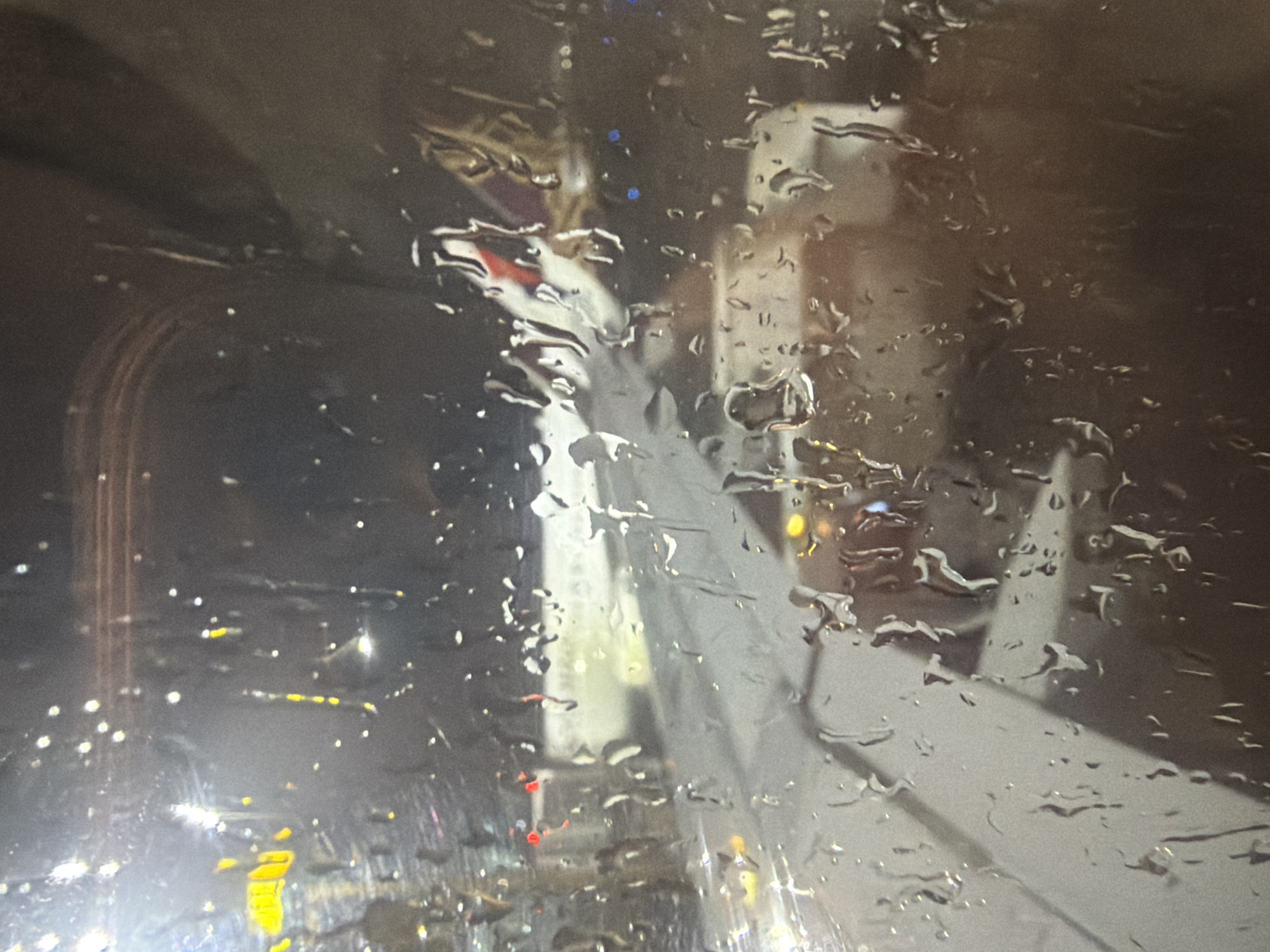 The image shows a view through an airplane window at night, with raindrops distorting the scene. An airplane wing is visible in the foreground, partially obscured by the water droplets. In the background, another airplane is visible, with its tail section showing a purple and red emblem. Lights from the airport or city are visible in the distance. The overall impression is of a rainy night at an airport, possibly Delhi, India, given the context provided.