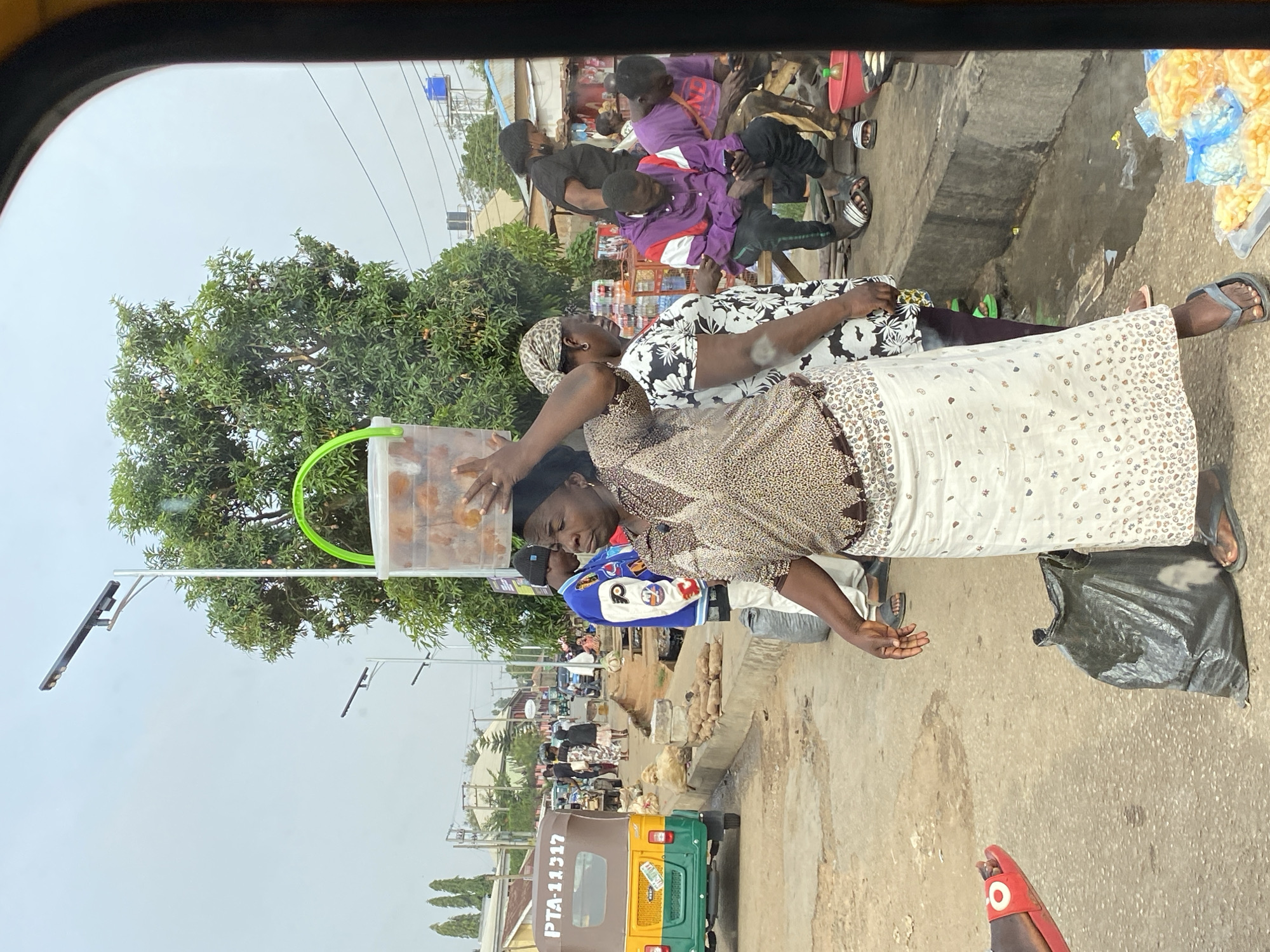 The image shows a street scene in Bukuru, Nigeria. An African woman wearing a patterned top and a white skirt with small designs is selling snacks from a clear plastic bucket on her head. She appears to be interacting with a young man wearing a blue jacket with a white sleeve and a black cap, who is standing on the sidewalk. Other people, including a woman in a black and white patterned dress, are visible in the background. Some people are sitting on what appears to be a roadside platform, possibly engaged in eating. A green and yellow auto-rickshaw is parked on the side of the road with the license plate number "PTA-11317" visible. In the background, there is a street lined with buildings and trees, and streetlights are present. The ground is paved. The image is taken from inside a vehicle, looking out.
