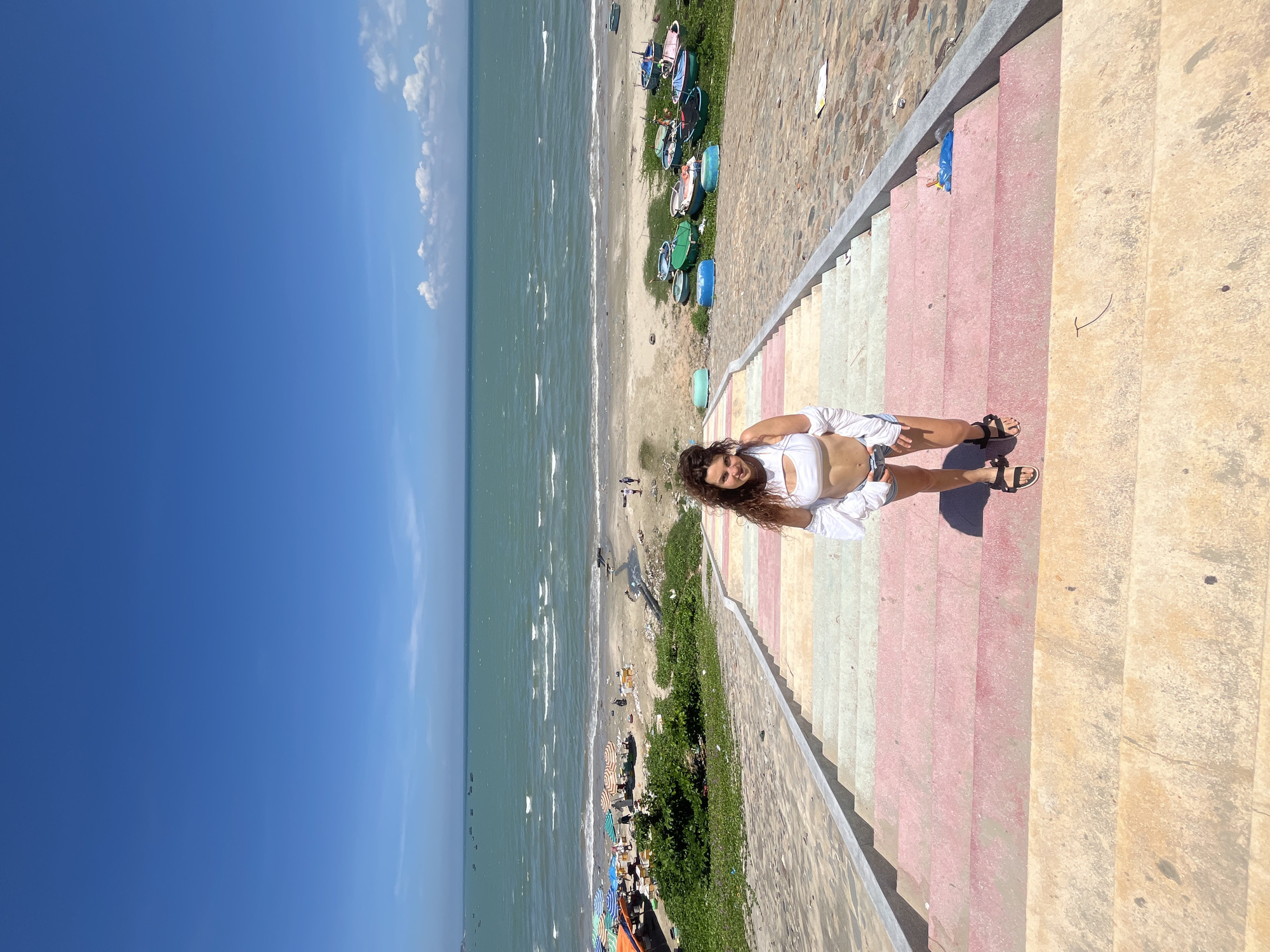 A young woman stands on a brightly colored staircase, smiling at the camera. The staircase is made of stone and painted in alternating stripes of yellow, pink, and light green. The woman is wearing a white crop top, denim shorts, and black sandals. Behind her, a beach scene unfolds. The turquoise ocean stretches to the horizon, meeting a clear blue sky dotted with a few white clouds. The sandy beach is lined with colorful umbrellas and small boats. The overall scene suggests a sunny day in Mũi Né, Vietnam, a popular coastal town known for its beaches and unique landscapes.