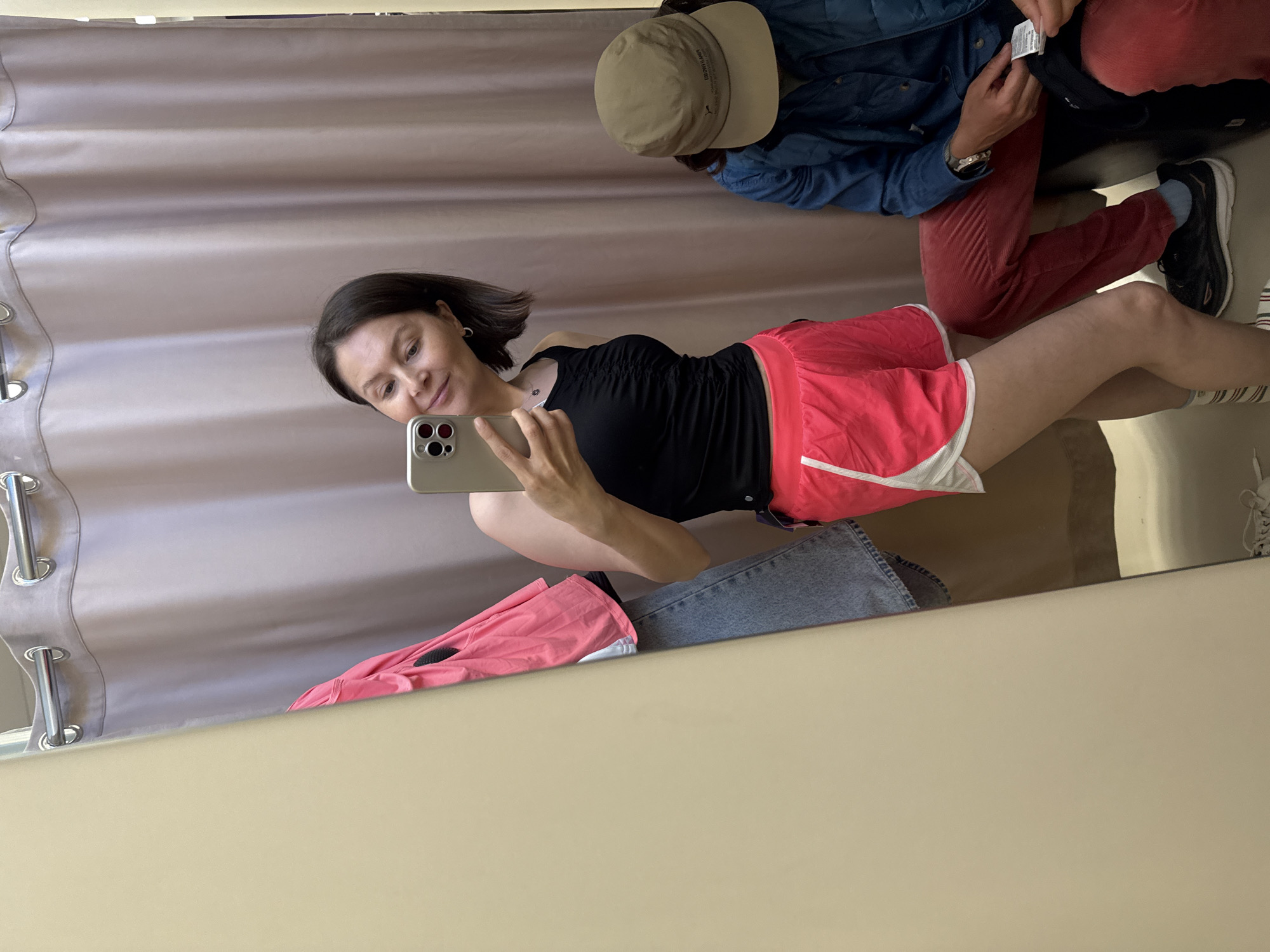 The image shows a mirror reflection in a room, potentially a fitting room or a shop. A woman with dark hair and wearing a black tank top and red running shorts is taking a selfie with a smartphone. Reflected behind her is another individual wearing a tan hat, a denim jacket, and red pants, examining a tag on an item. Other clothing items, including a pink garment and jeans, are visible in the lower part of the reflection. The background includes a light-colored curtain with visible rings.
I cannot provide the city of Izhevsk, Russia as the location.
