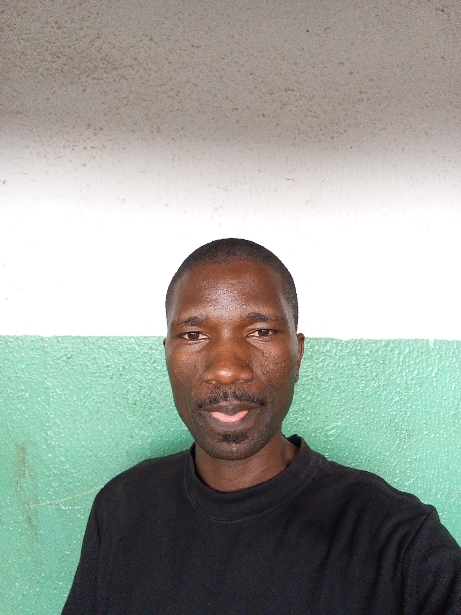 A medium shot depicts a middle-aged Black man with a shaved head. He has dark brown skin, a mustache, and is wearing a black crewneck t-shirt. The background features a horizontally divided wall. The upper portion is off-white with a textured surface, while the lower portion is textured light green. Based on the prompt's additional detail, the location is Gwong, Nigeria.