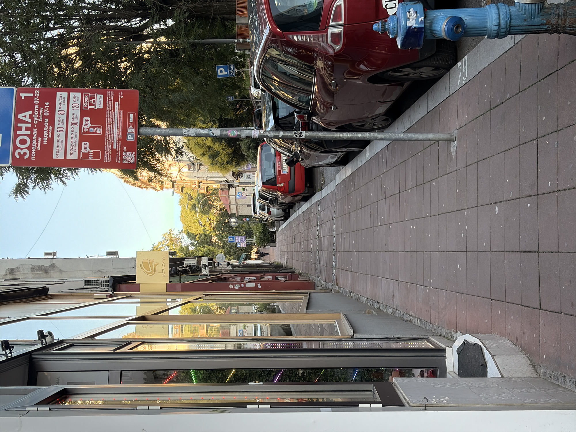 This image depicts a street scene in Belgrade, Serbia. On the left, a storefront with large windows displays colorful lights. A sidewalk paved with red-brown tiles runs alongside the buildings. A prominent red parking sign with Cyrillic text is visible, indicating parking zone information and payment methods. Several cars are parked along the street. A blue fire hydrant stands on the sidewalk near the foreground. The overall environment appears to be a commercial area in a city, with buildings and trees lining the street. The sky is clear and blue, suggesting it is daytime.
