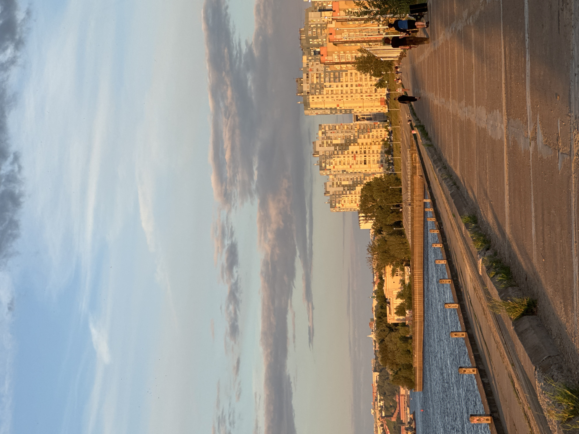 A horizontal, wide-angle, eye-level shot shows a cityscape and a waterfront promenade under a soft, hazy sky. The sky is a pale blue with streaks of light grey and pink clouds. In the background, numerous apartment buildings are visible, with their windows reflecting the warm, late afternoon light. Trees and some scattered foliage line the waterfront and intersperse the buildings.
The foreground features a wide, paved walkway that appears to be a promenade or embankment. The surface of the walkway is textured and shows some signs of wear, with white lines marking sections. The promenade is bordered by a concrete barrier on the side facing the water. Along the water's edge, there is a series of small, evenly spaced concrete posts. The water itself is a deep blue, calm, and reflects the sky.
Several people are present on the promenade, walking or standing. Some are silhouetted against the bright light. One person is seated, and another is walking away from the camera. The overall mood is peaceful and serene, suggesting a leisurely late afternoon or early evening. The low angle of the sun indicates it is likely near sunset, casting long shadows and bathing the scene in a warm, golden light. The location is identified as Nizhny Novgorod, Russia, which is known for its waterfront areas along the Volga River. There is no visible text in the image.