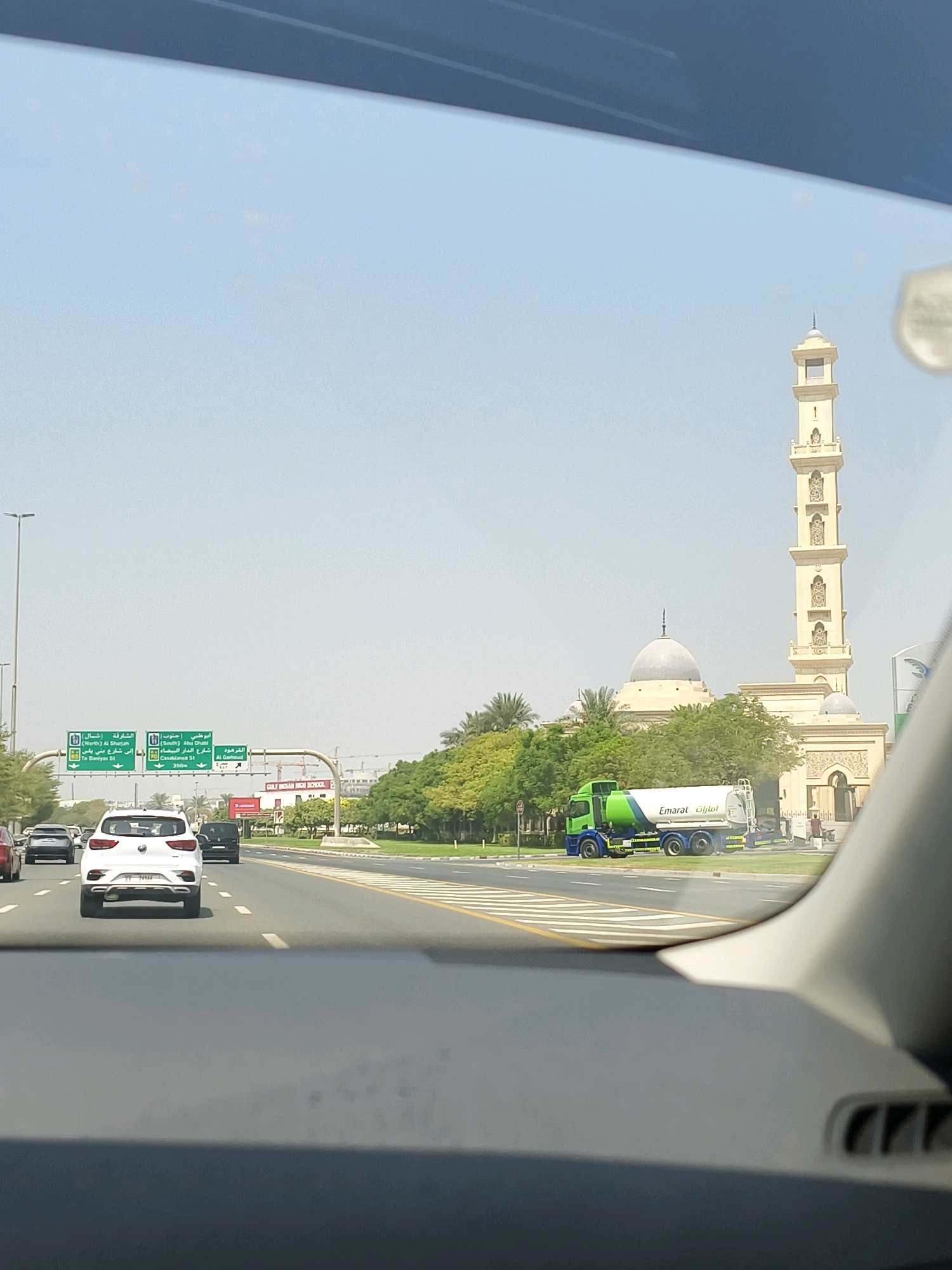 The image captures a view from inside a vehicle traveling on a multi-lane highway in Dubai, United Arab Emirates. The sky is a clear, pale blue, suggesting a sunny day. On the right side of the road, a prominent mosque with a tall, slender minaret and a large dome is visible, constructed with light-colored stone. In front of the mosque, lush green trees and landscaping line the roadside.
Several cars are driving on the highway, including a white SUV in the foreground. A green and white tanker truck with "Emarat" branding is also present on the road. Overhead green highway signs display directions in both Arabic and English, indicating directions to "Al Shuja (North)", "Abu Dhabi (South)", and "Al Ghaaf (East)". One sign specifically points to "Casablanca St" and "To Danyas St". In the background, beyond the trees, a building identified as "GULF INDIAN HIGH SCHOOL" is visible. Construction cranes are also present in the distance, indicating ongoing development in the area. The road surface is paved and marked with white and yellow lines. The interior of the vehicle from which the photo is taken is partially visible in the foreground, with the dashboard and windshield frame.
