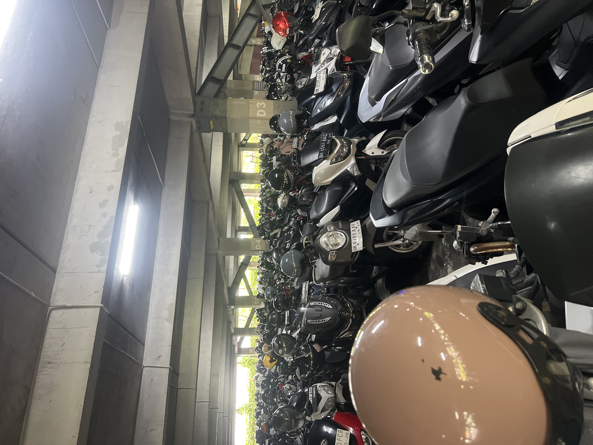 The image depicts a dense parking lot for motorcycles in Kuta, Indonesia. Rows of motorcycles are tightly packed between concrete pillars and structural supports, extending deep into the frame. A brown helmet is prominently featured in the foreground, partially obstructing the view. Signage with section designations "D3" and "D4" is visible amongst the rows of bikes. License plates are visible on many of the motorcycles. Fluorescent lighting is present in the ceiling.