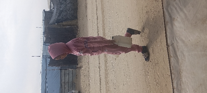 A person walks right-to-left on a dirt path in Khan Yunis, Palestinian Territory. The individual wears a pink hooded tracksuit with dark stripes on the sleeves and a white graphic on the chest, along with dark sandals. They carry a light-colored plastic jerry can by its handle in their right hand. The background features several makeshift shelters constructed from corrugated metal panels and dark fabric tarpaulins. The sky is overcast.