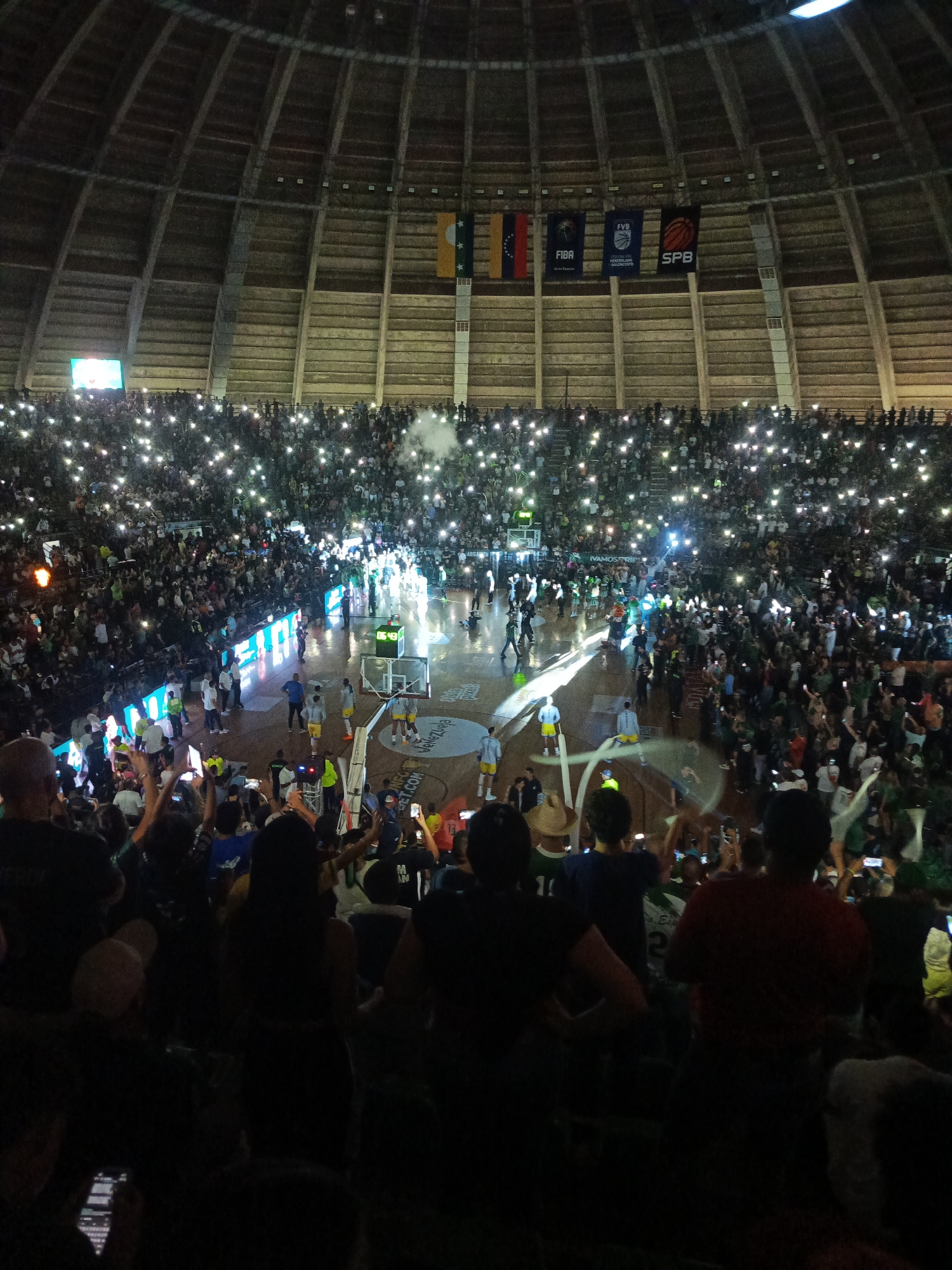 This is a medium-angle, indoor shot of a basketball game in a large, circular arena. The arena is filled with spectators holding up their phone flashlights, creating a starry effect in the stands. The court is lit, and players in yellow and white uniforms are visible on the court, along with officials and staff. Banners hang from the ceiling, including flags and logos such as FIBA and SPB. The scene appears to be from a professional basketball game, likely within the city of La Asuncion, Venezuela, given the presence of the SPB (Superliga Profesional de Baloncesto) logo. The overall atmosphere is energetic and celebratory.