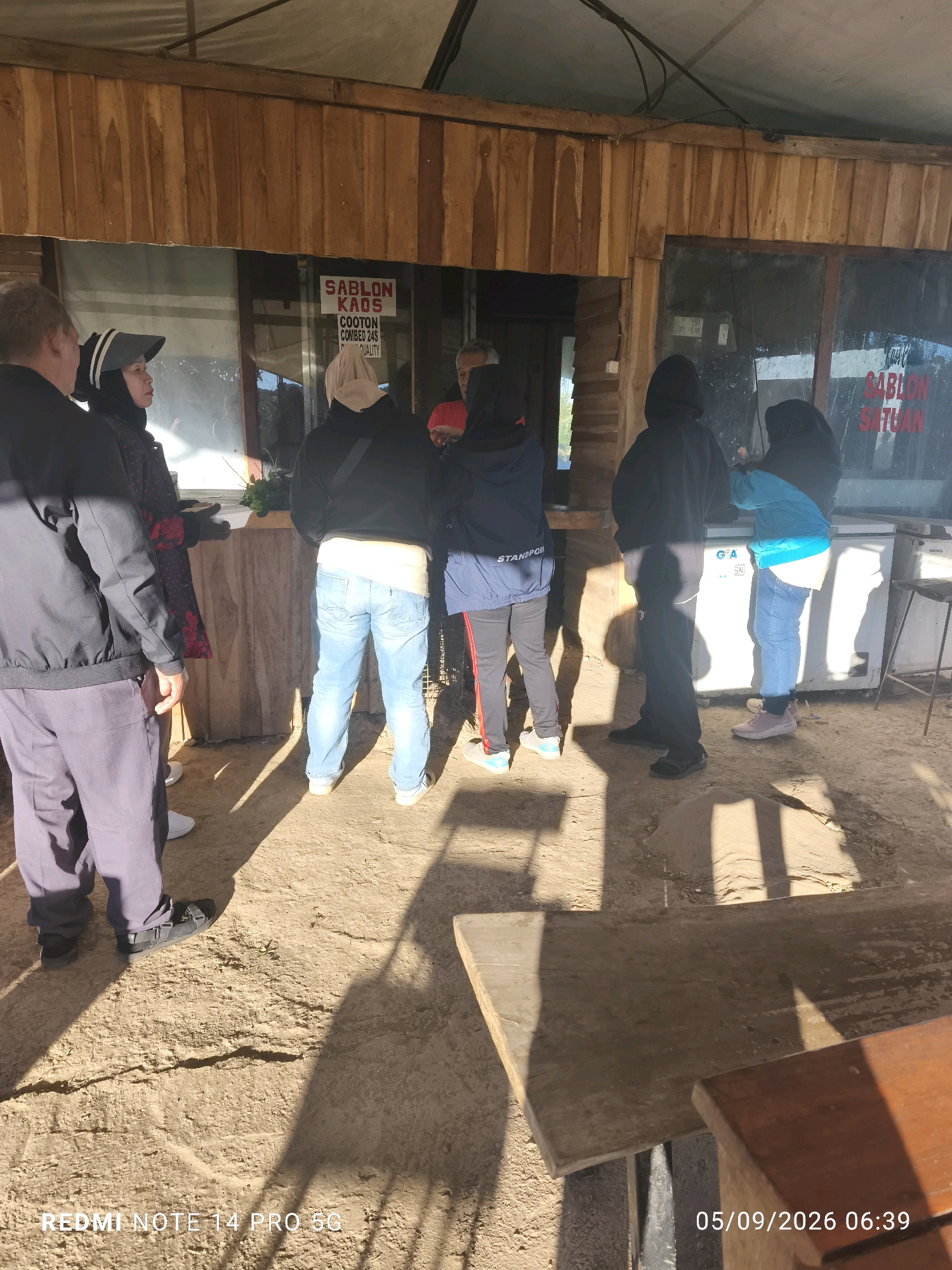 A group of people stand outside a wooden structure with large windows. A sign on the window reads "SABLON KAOS COOTON COMBED 24S QUALITY". A person wearing a blue jacket and jeans stands at a white refrigerator. The ground is sandy and dry. The scene appears to be outdoors, possibly at a marketplace or shop, in the city of Rancabali, Indonesia.