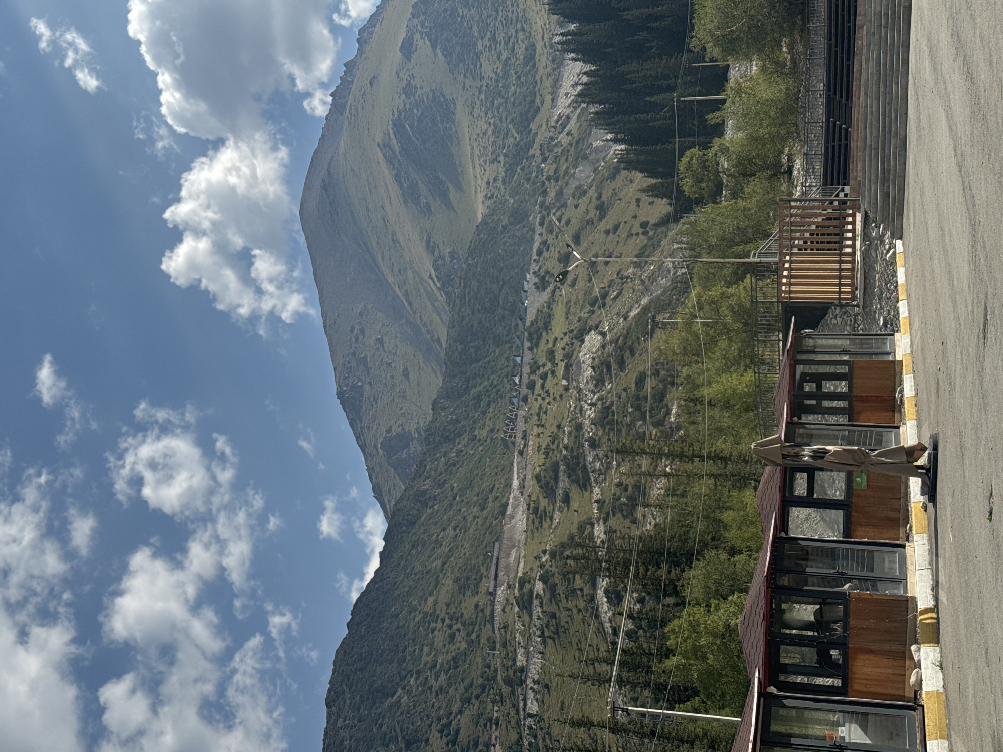 In the city of Unknown, Kyrgyzstan, a mountainous landscape dominates the scene under a partly cloudy blue sky. In the foreground, a row of small, covered outdoor dining or seating areas with wooden siding and glass windows is visible. An umbrella stand is positioned in front of one of the structures. Adjacent to these, a set of concrete stairs ascends towards a wooden fence and an empty amphitheater-like seating area. Further into the background, a road winds its way up the green, rocky slopes of the mountains, dotted with sparse vegetation and trees. A line of dark green coniferous trees marks the base of the steeper mountain slopes. The overall impression is of a recreational area nestled within a rugged natural environment.