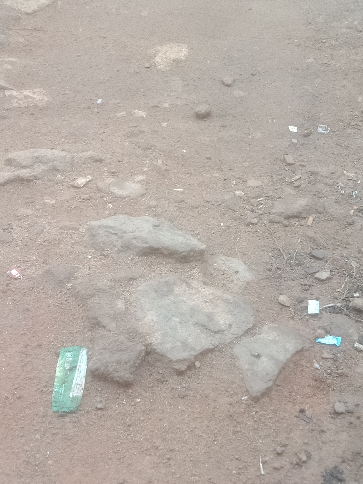 An overhead, eye-level view of a dirt and rock-strewn ground. The ground is mostly a light brown color with varying shades of grey rocks scattered throughout. There are several pieces of trash visible on the ground. In the bottom left corner, there is a green and white wrapper. Further up and to the right, there are several small pieces of white and blue paper. The scene appears to be outdoors in a natural or undeveloped area.
