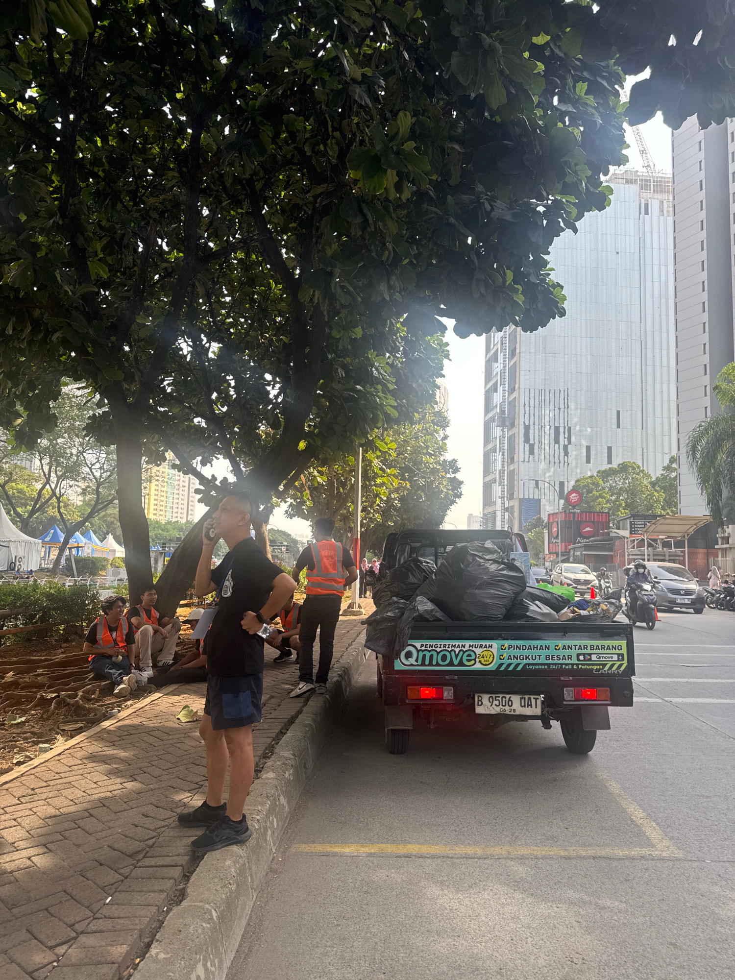 A man in a black t-shirt and blue shorts stands on a sidewalk in front of a pickup truck loaded with black trash bags. The truck has a license plate that reads "B 9506 OAT". Several men wearing orange safety vests are sitting on the ground to the left of the truck, under a large tree. In the background, tall buildings and construction cranes are visible. The scene appears to be on a street in South Jakarta, Indonesia, with a sidewalk and road present.