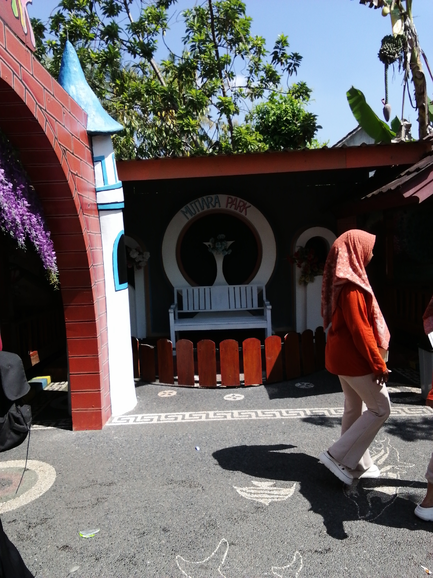 In Tegalwangi, Indonesia, the image captures an outdoor scene featuring a distinctive architectural element resembling a castle tower, painted in white with blue accents and topped with a blue conical roof. Adjacent to this structure is a red brick archway adorned with hanging purple flowers. The ground is paved with asphalt and decorated with white chalk drawings. A brown wooden fence encloses a section of the area. In the background, a dark wall displays a circular motif with the text "MUTIARA PARK" and a white bench is visible. A woman, dressed in an orange hijab and shirt with light-colored pants and white sneakers, walks across the pavement, casting a shadow. Another person, partially visible in the lower right, also wears light-colored pants and white shoes. The environment is filled with lush green trees and a clear blue sky.