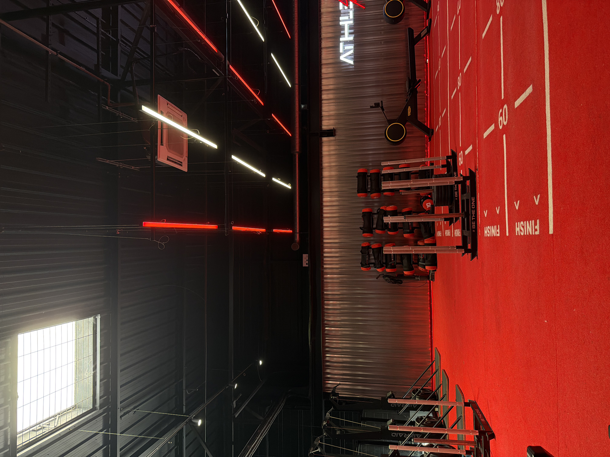 The image depicts the interior of a modern gym, identified as Athluz, located in Strasbourg, France. The environment is characterized by a vibrant red carpet flooring marked with white lines and numbers, suggesting a functional training or track area. Various exercise equipment is visible, including sleds, rowing machines, and weight bags. The ceiling is industrial black, featuring a skylight and a grid of red and white LED lights, along with a suspended air conditioning unit. The back wall is corrugated metal, illuminated by red lighting, and features the "ATHLUZ" logo in white neon. The overall scene conveys a high-energy, contemporary fitness space. There are no people visible in the image.
